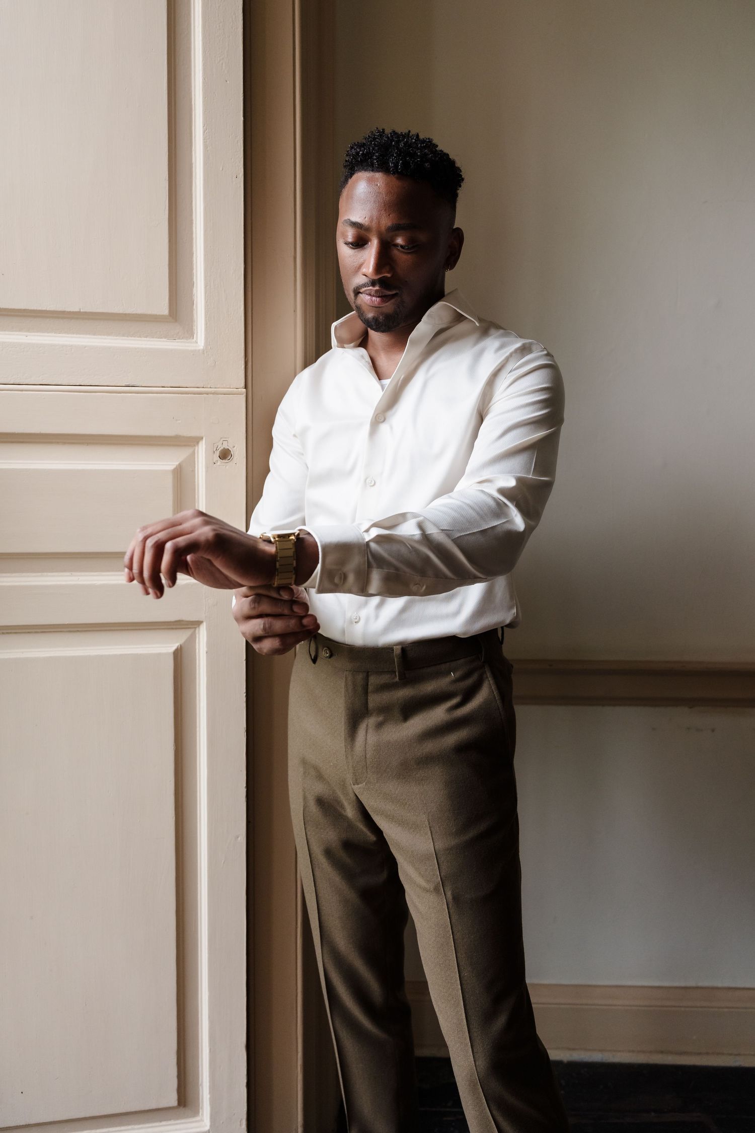 Stylish groom in white shirt and brown pants poses elegantly against a minimalist wall.