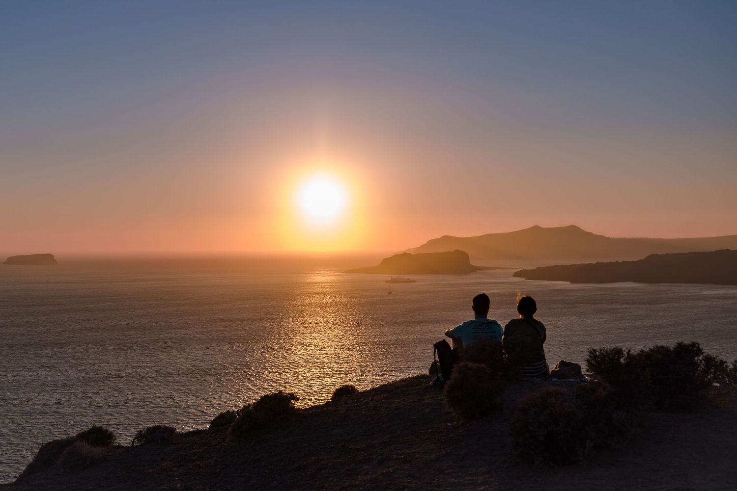 Two silhouettes sit on rocks watching a golden sunset over the ocean and mountains.