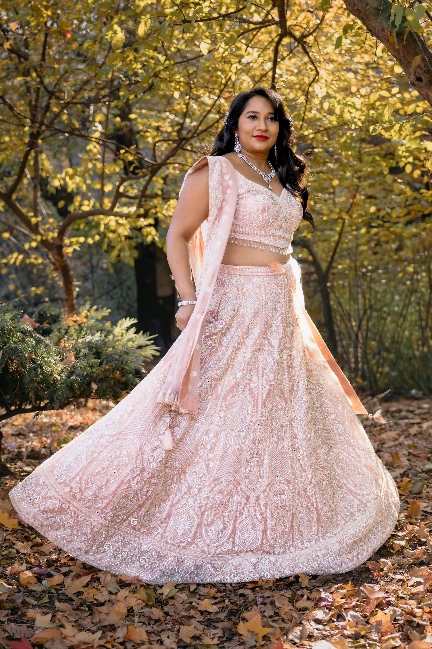 A pink lehenga with detailed embroidery twirls in an autumn forest setting.