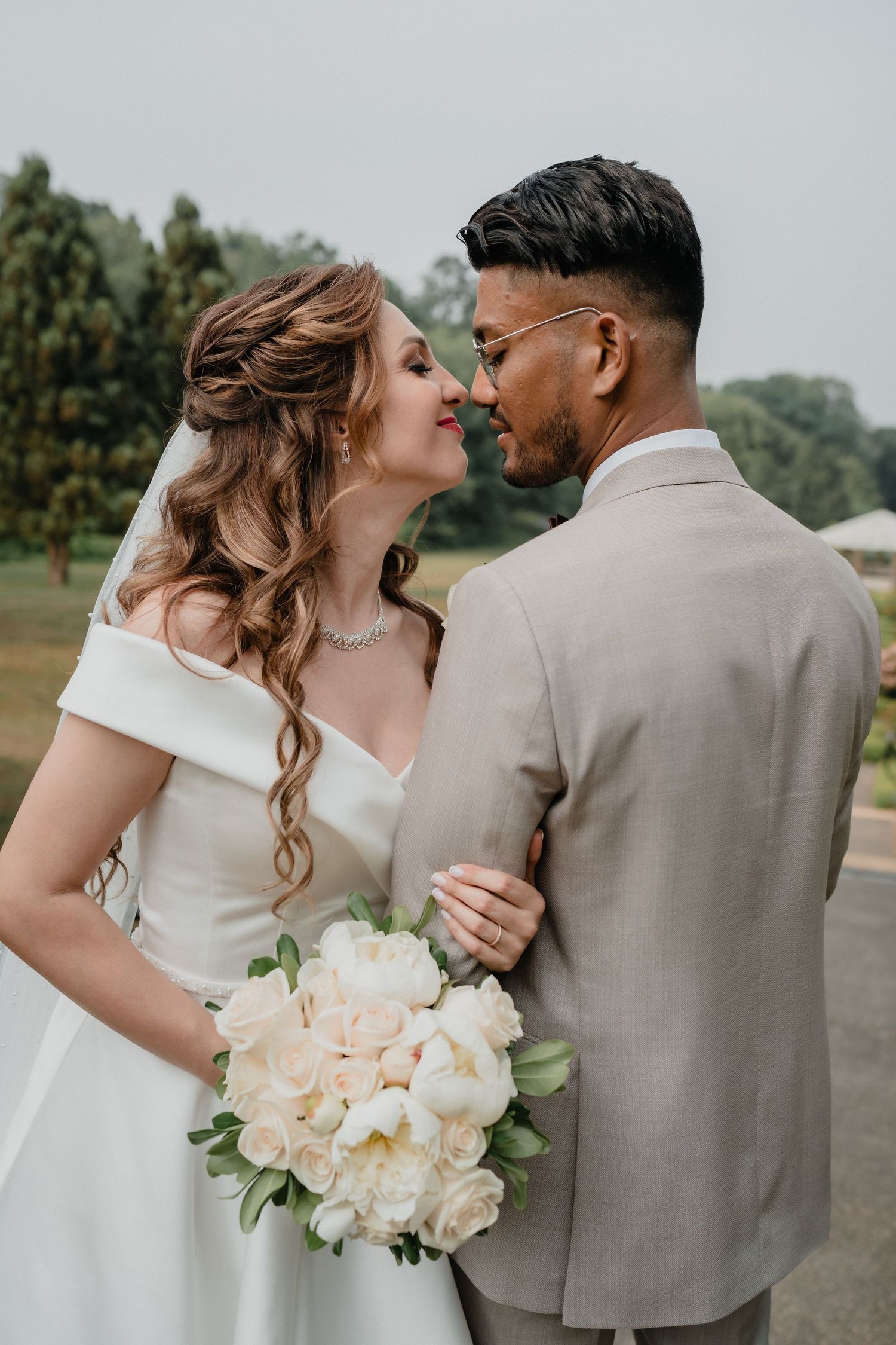 Romantic wedding couple embracing outdoors with white floral bouquet and beige suit.