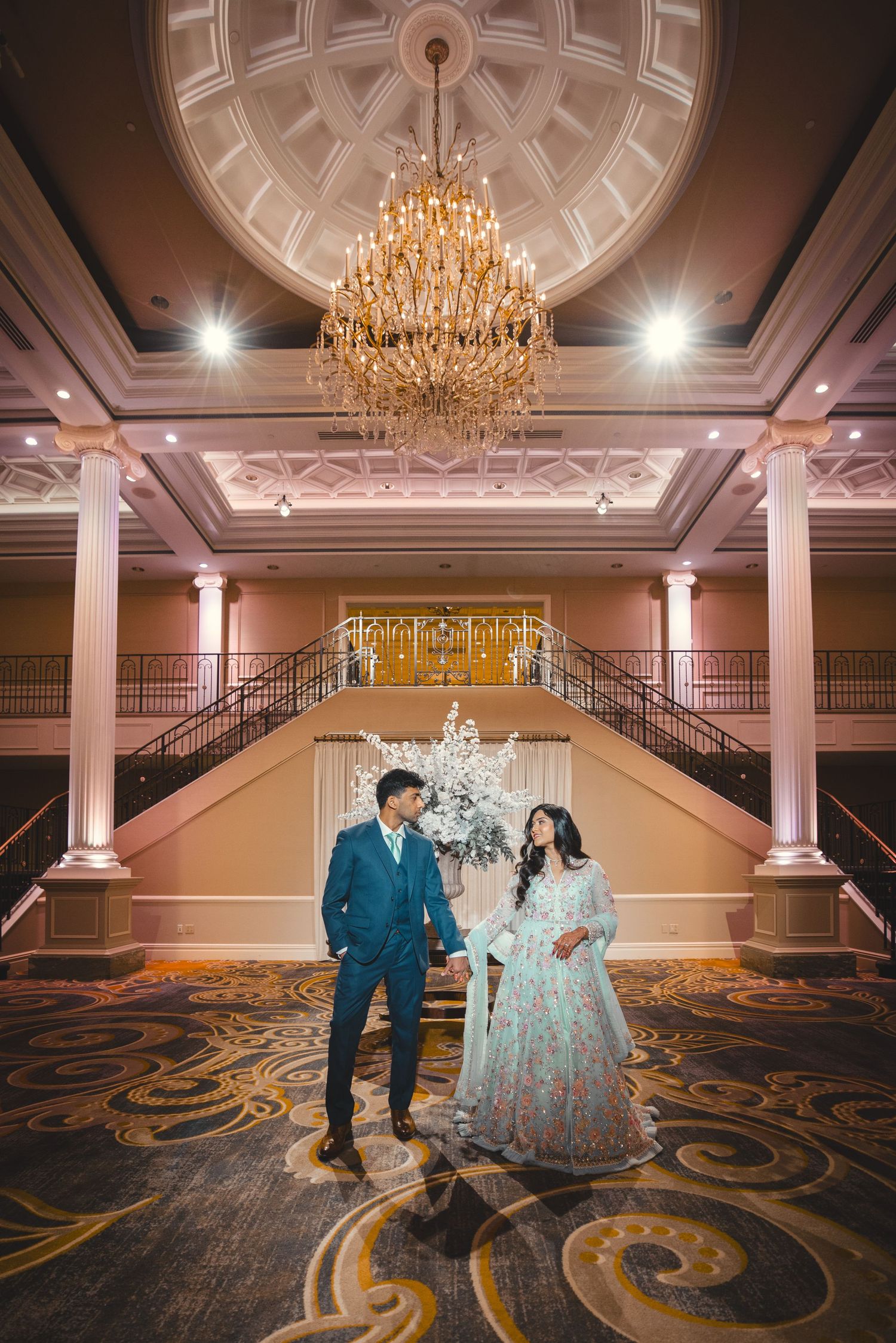 Grand ballroom with ornate chandeliers and decorative carpet features elegant celebration setup.