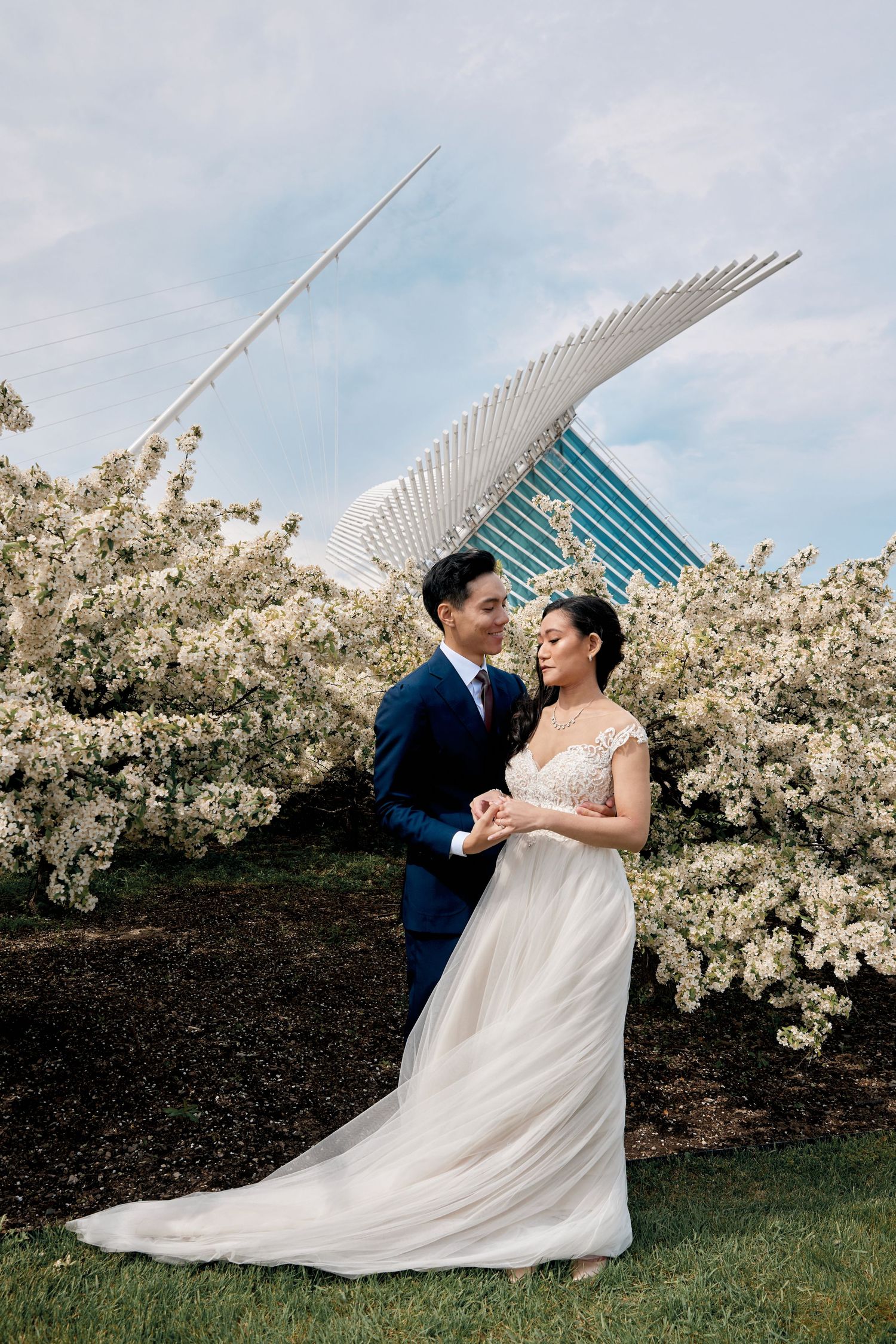 A couple poses among blooming cherry trees with modern architecture in background.