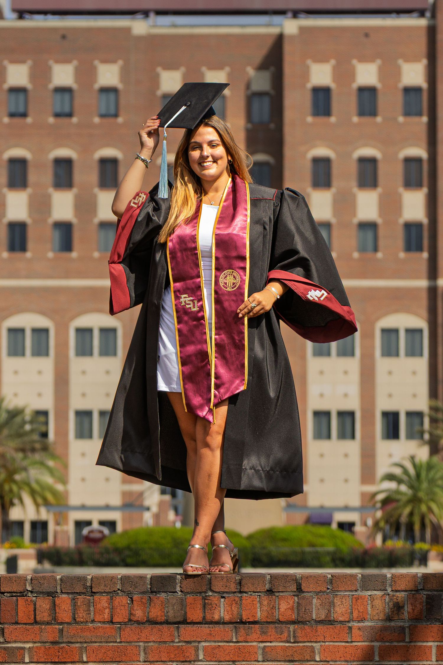 Graduate in cap and gown poses on brick wall for celebratory photos at university.