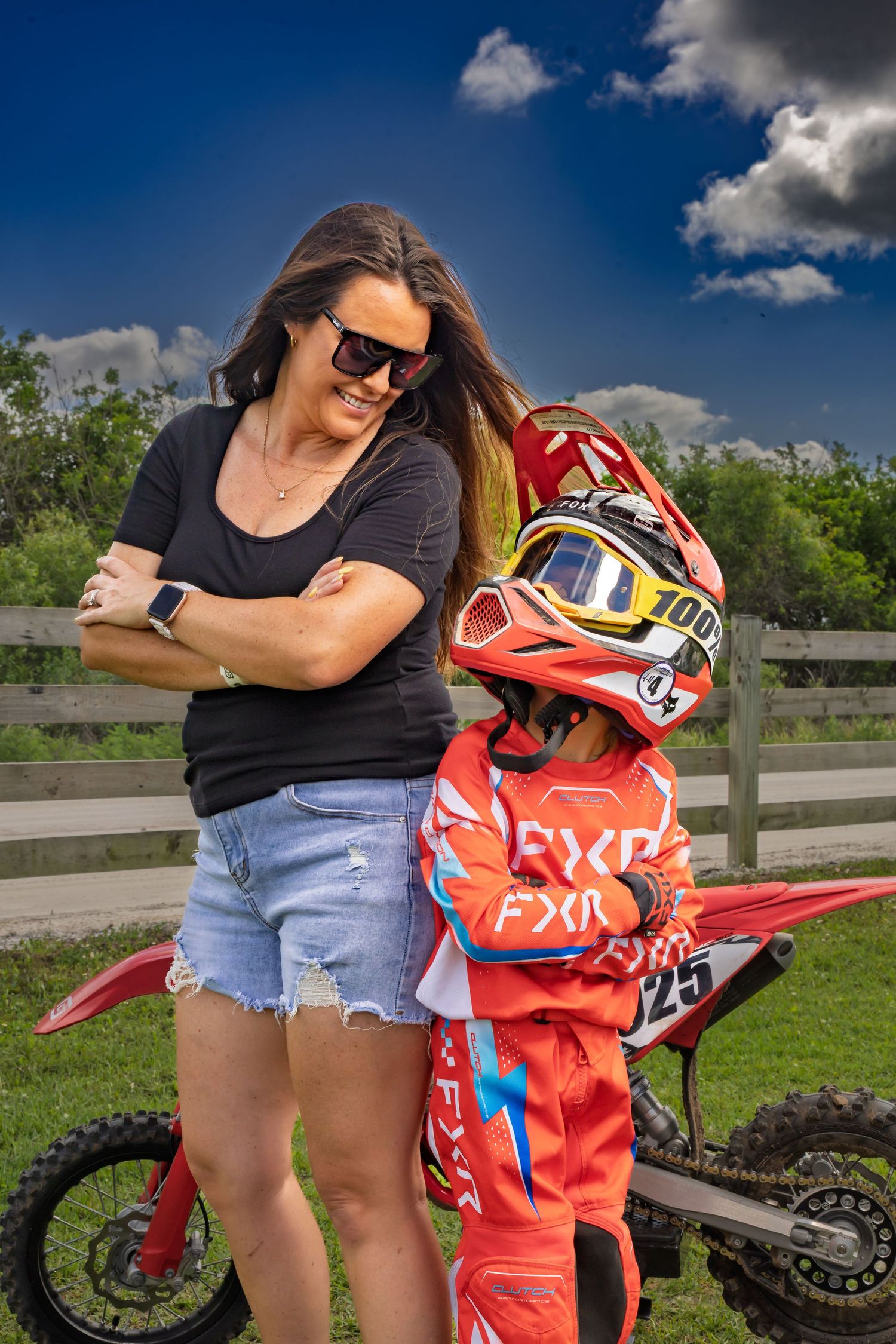 A woman in sunglasses and denim shorts stands proudly beside a young motocross rider in full FXR racing gear and helmet.
