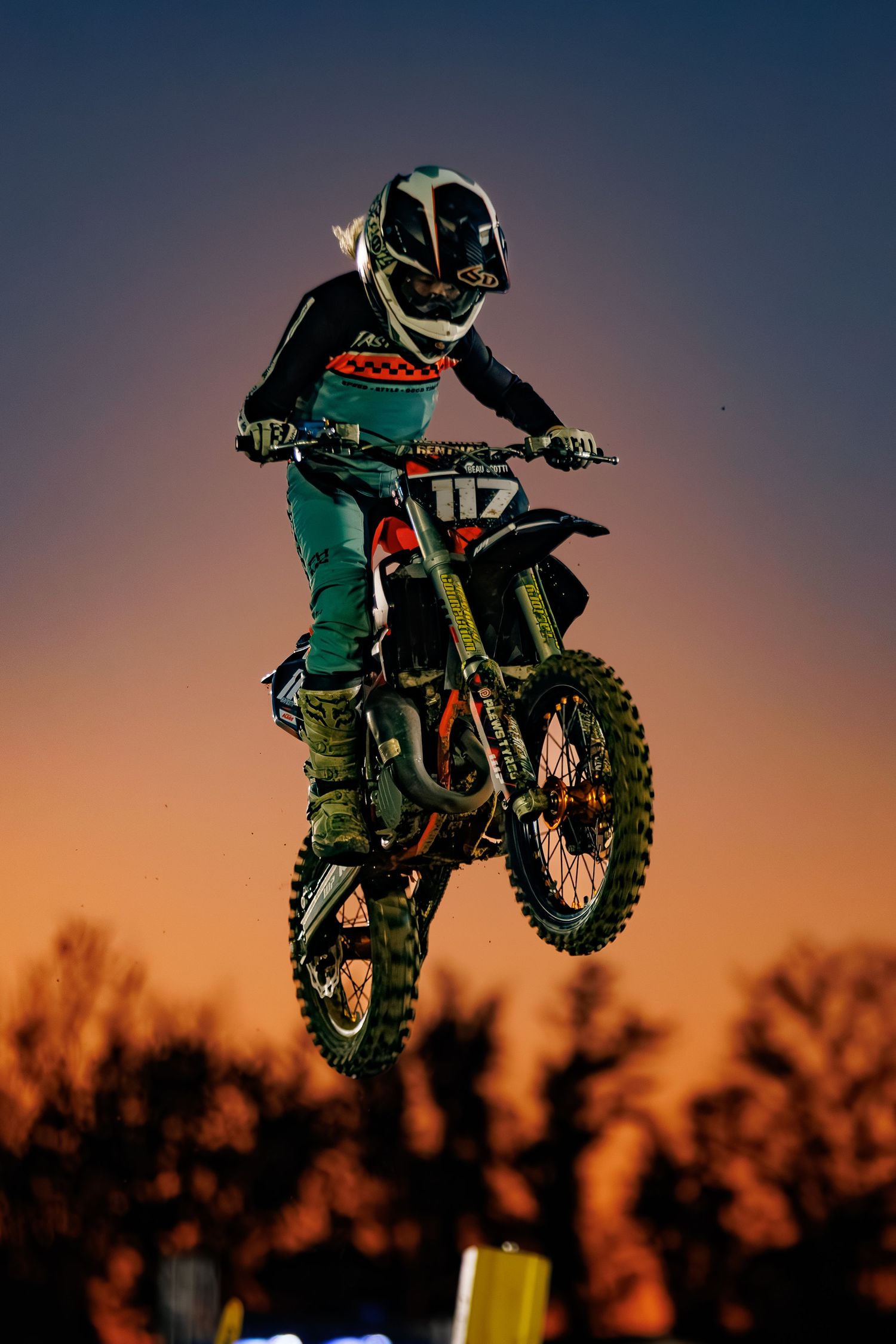 Motocross rider wearing teal gear launches into the air on a dirt bike against a vibrant orange sunset sky.