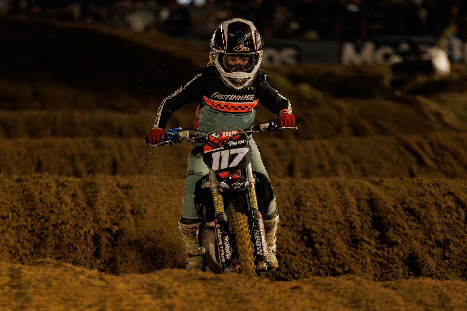 Motocross rider #117 racing on a dirt track at night under bright lights during a competitive event.