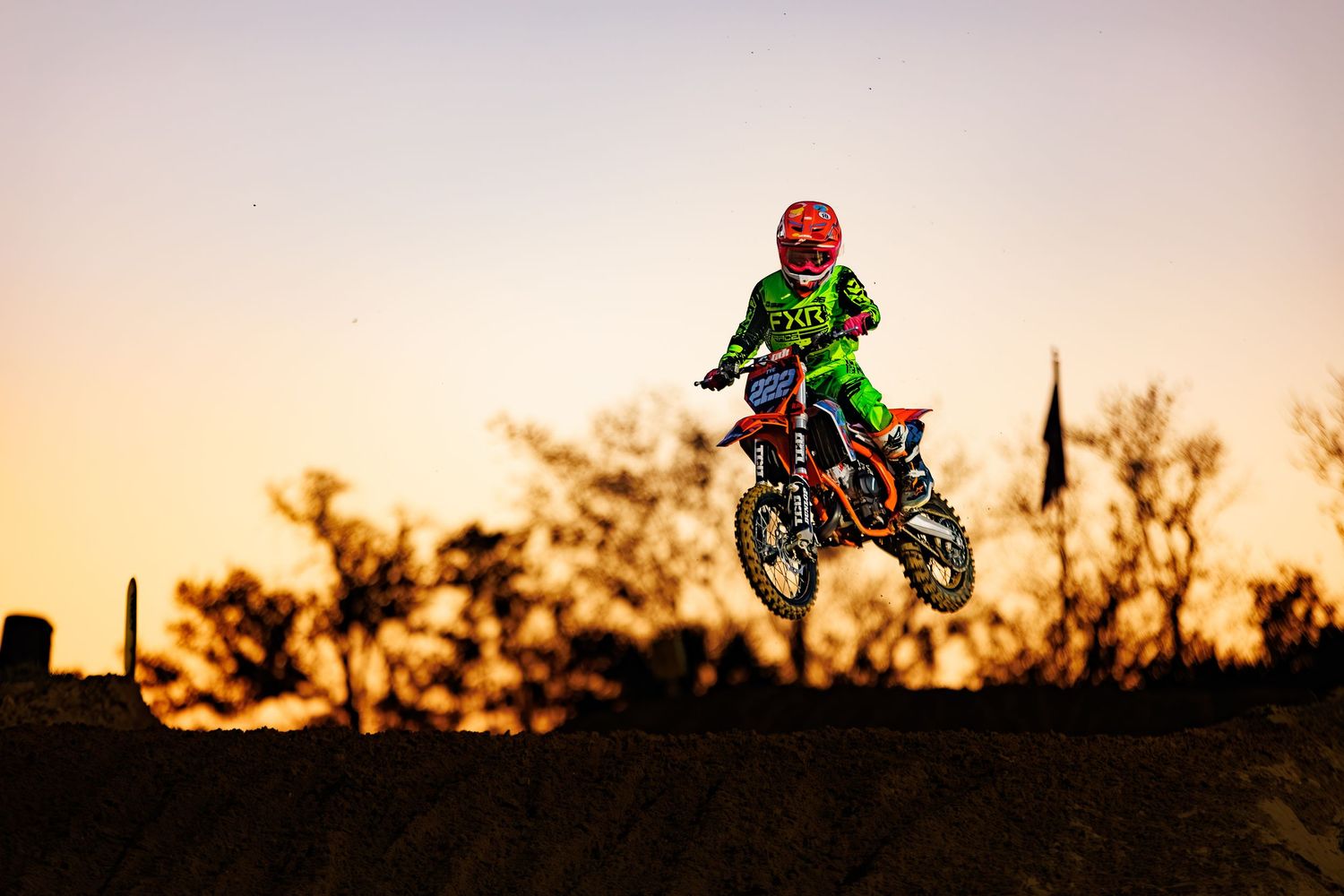 Young motocross rider in green gear jumps dirt bike against stunning golden sunset sky silhouette.