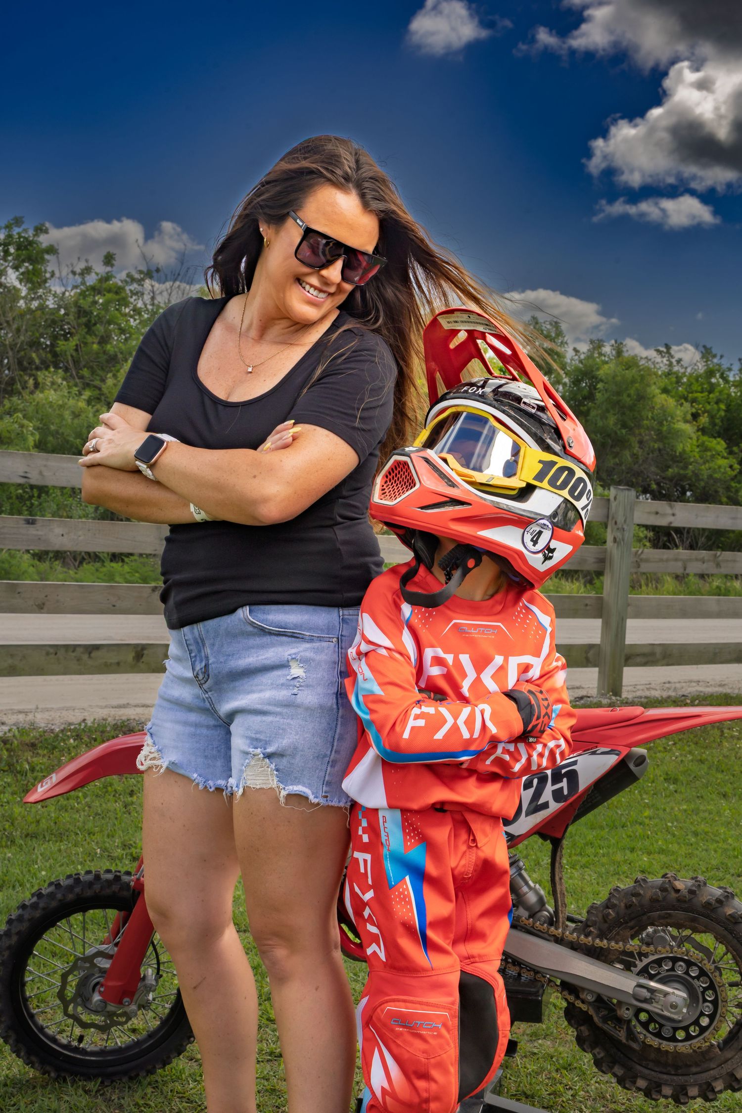 A woman in sunglasses and denim shorts stands with a child in FXR motocross gear and red helmet beside a dirt bike.
