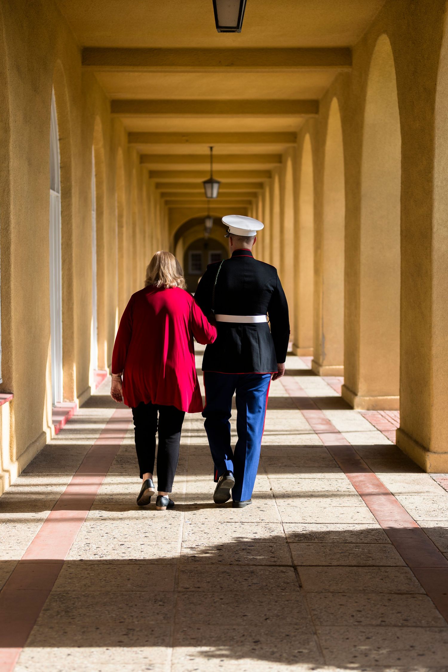 A couple walks through a sunlit colonnade with repeating arches and shadows creating a geometric pattern.