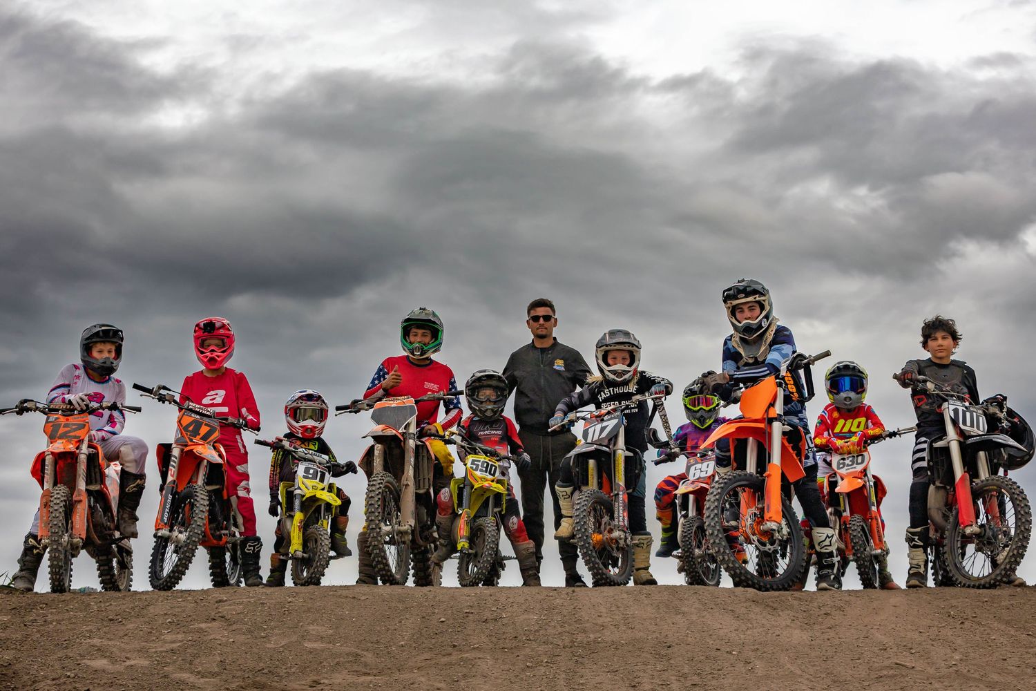 A group of young motocross riders with dirt bikes lined up on a track under dramatic cloudy skies with an adult coach.