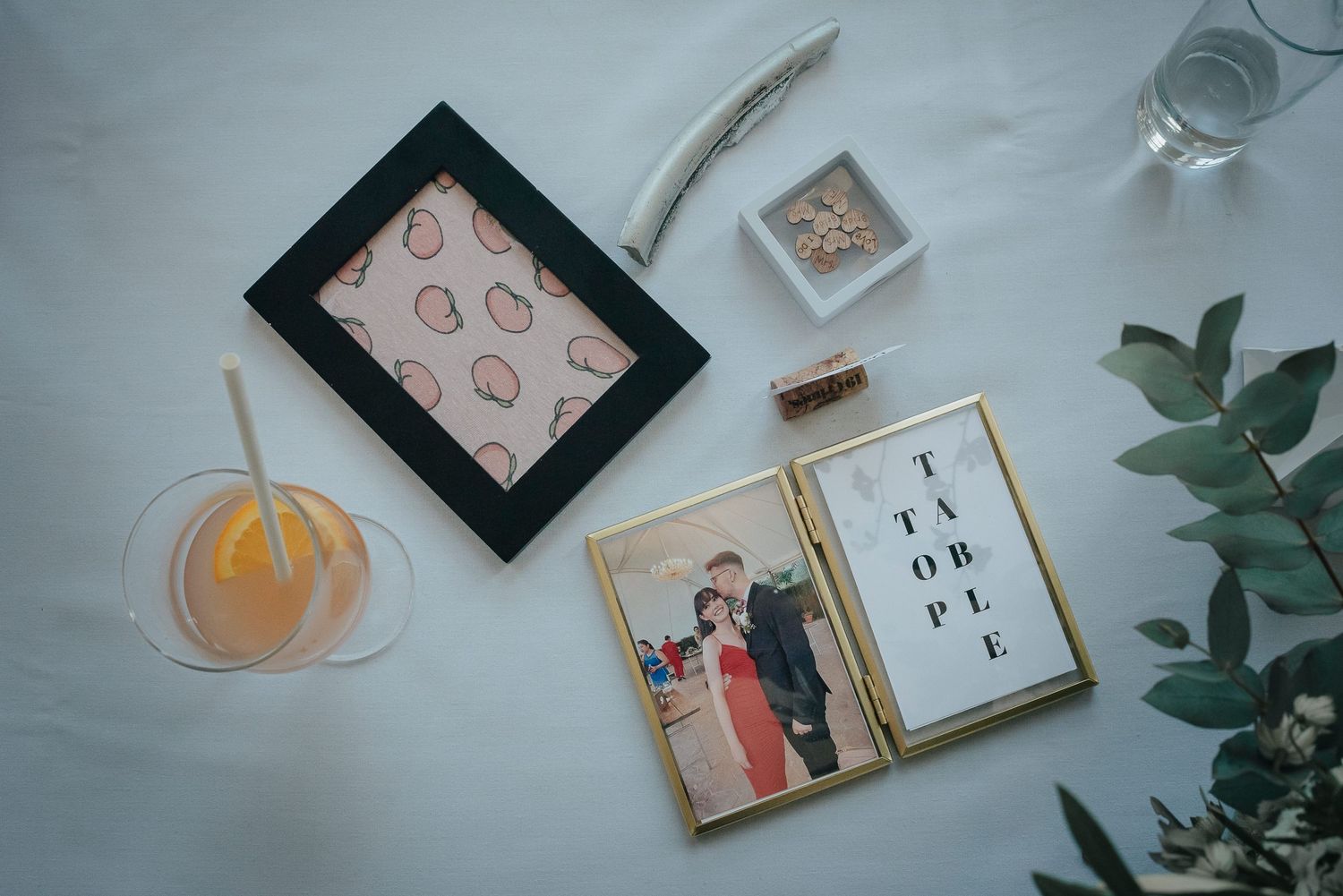 Flat lay of wedding stationery and decorative items on white surface with orange drink.