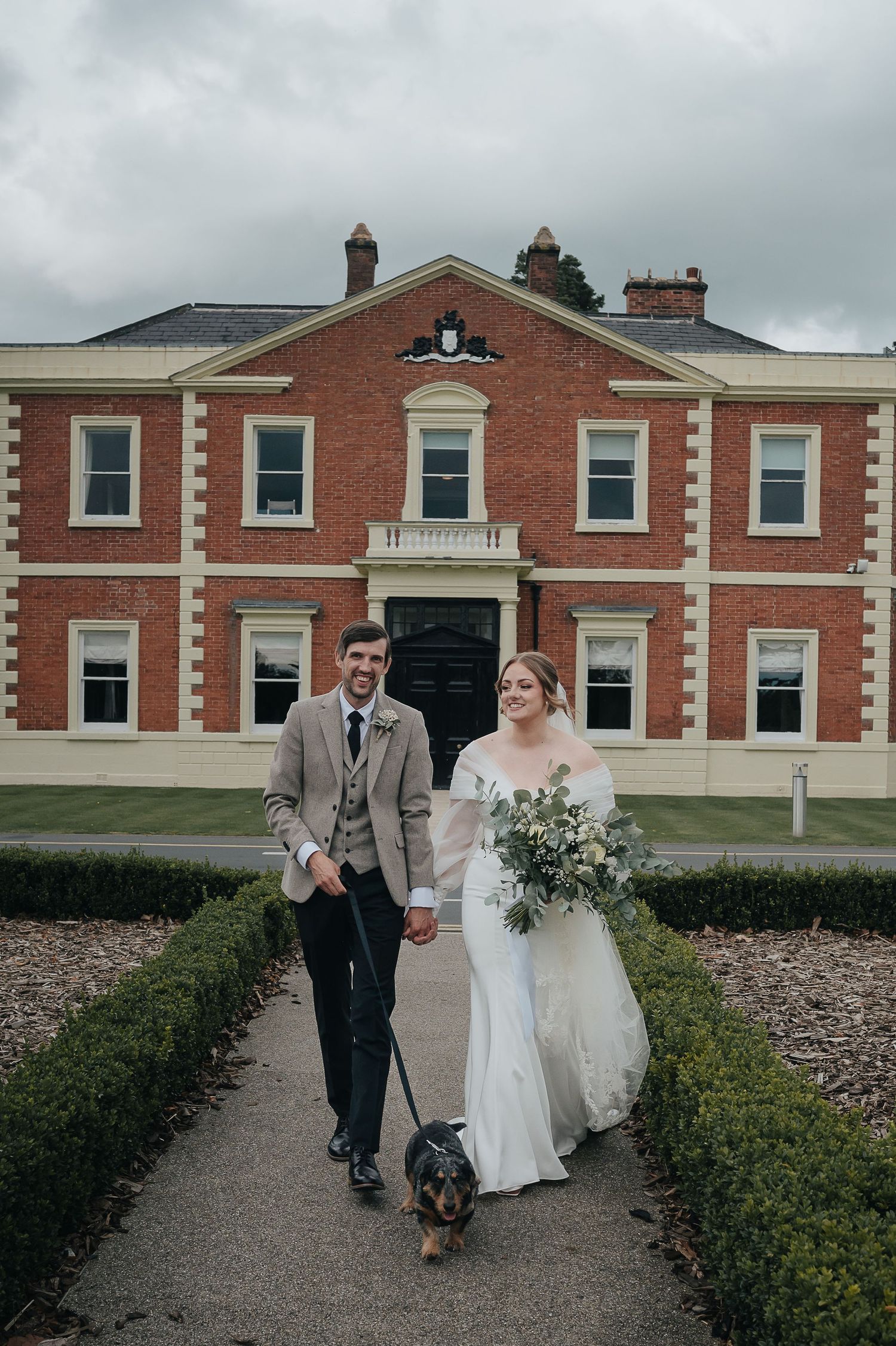 A couple walks with their dog on a path in front of a grand red brick mansion.