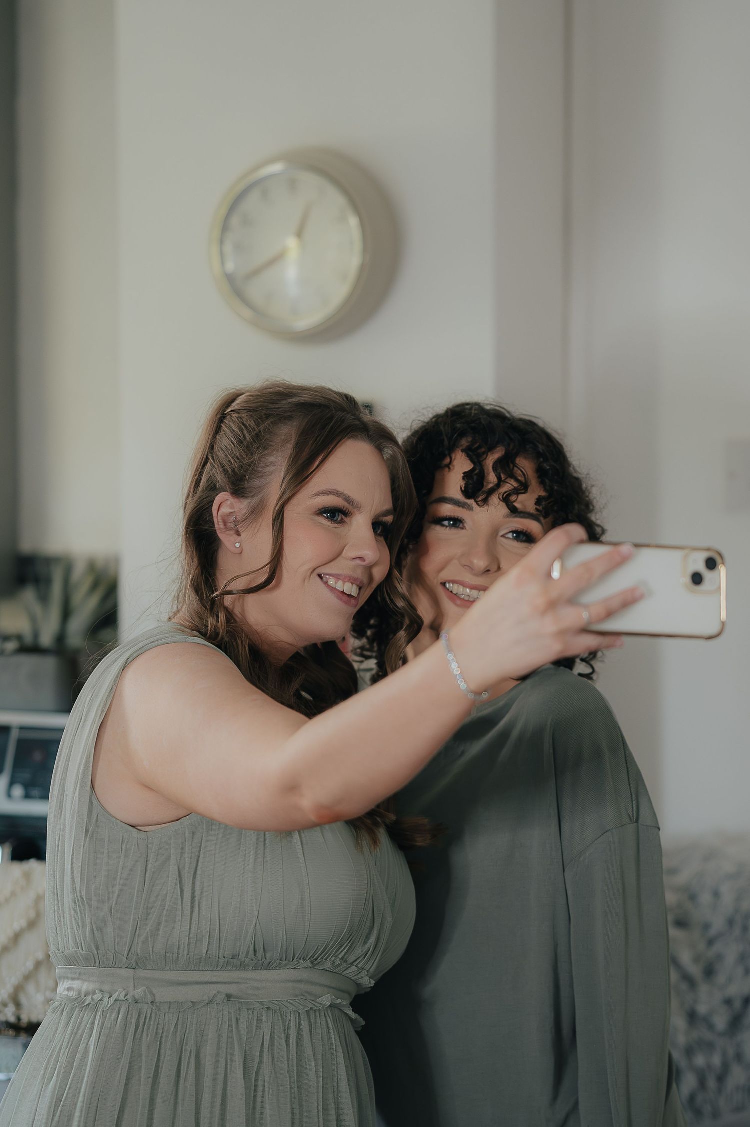 Two people in sage green dresses taking a selfie together by a wall clock.