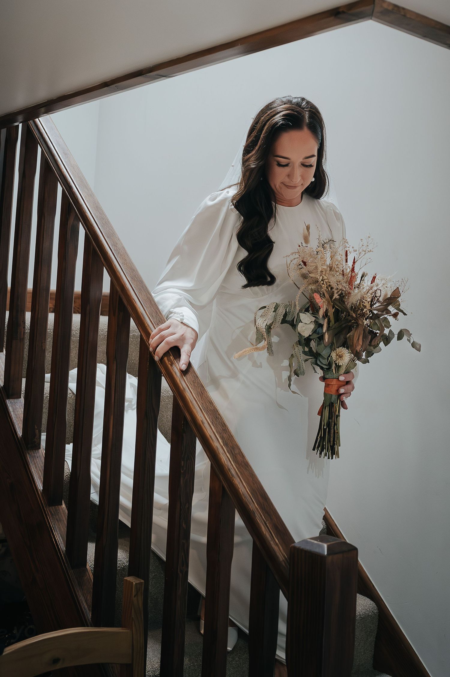 A bride descends a wooden staircase while holding a rustic bouquet of dried flowers.