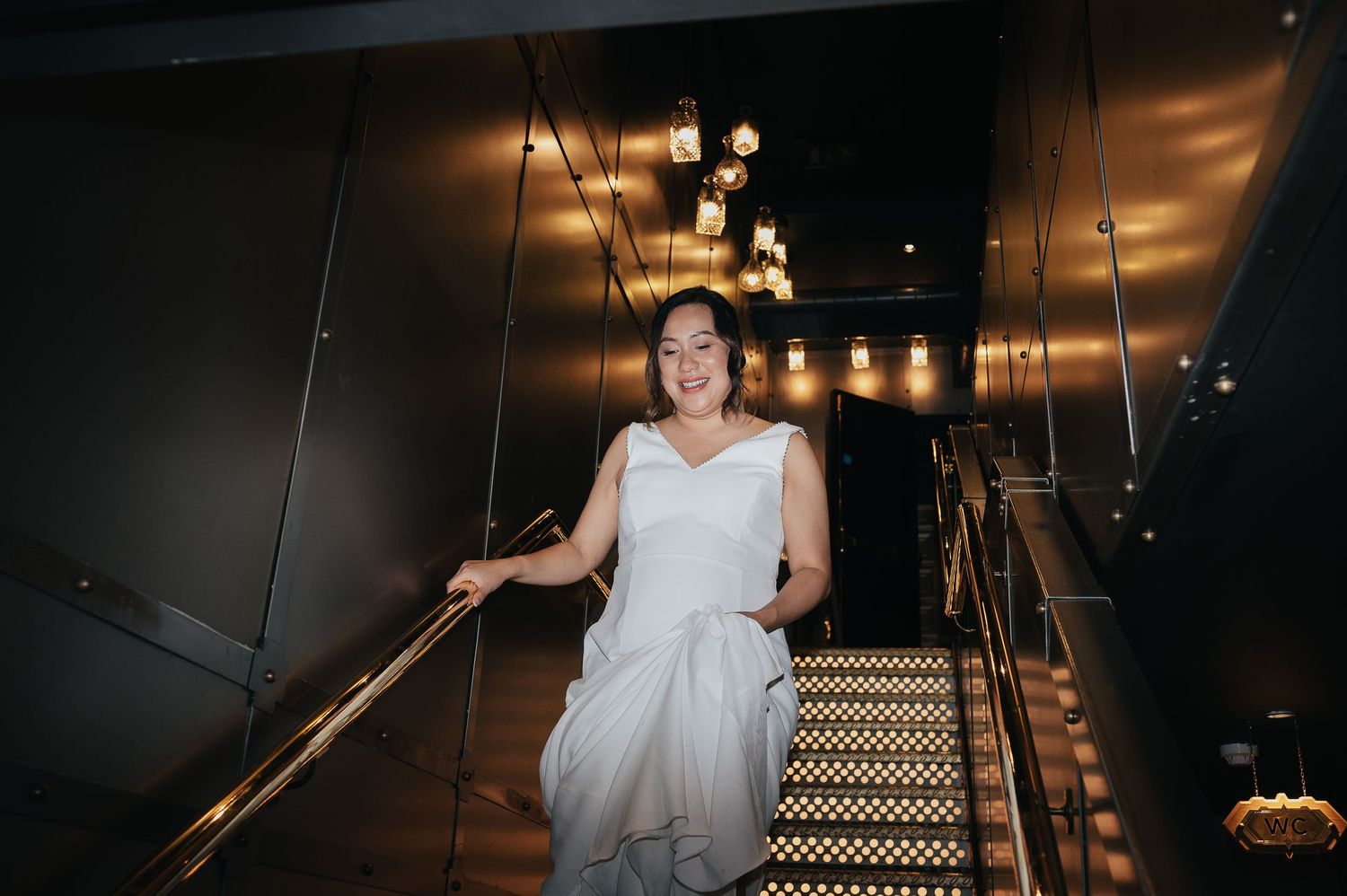 Bride in wedding dress descending modern staircase with illuminated ceiling.