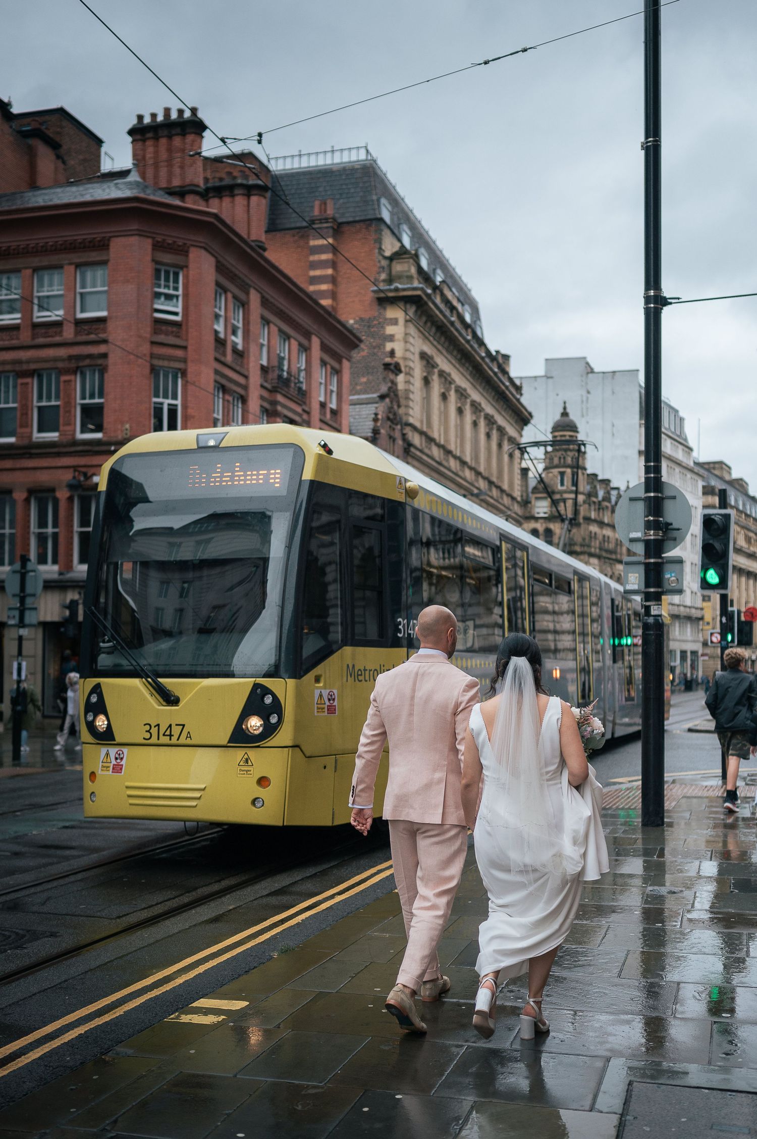 A couple in wedding attire walks past a yellow tram on a rainy city street.