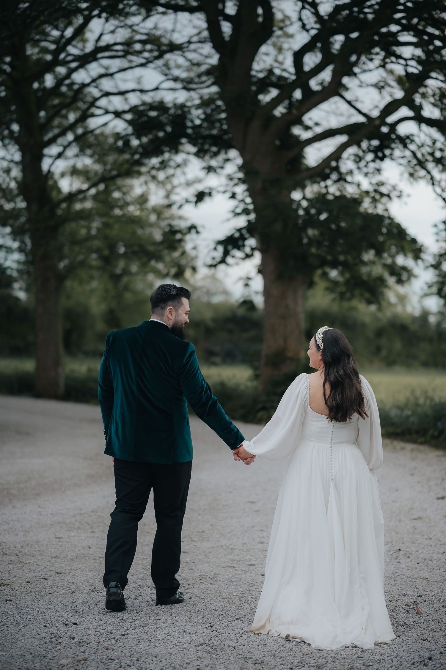 Couple in wedding attire walking hand in hand down a tree-lined path.
