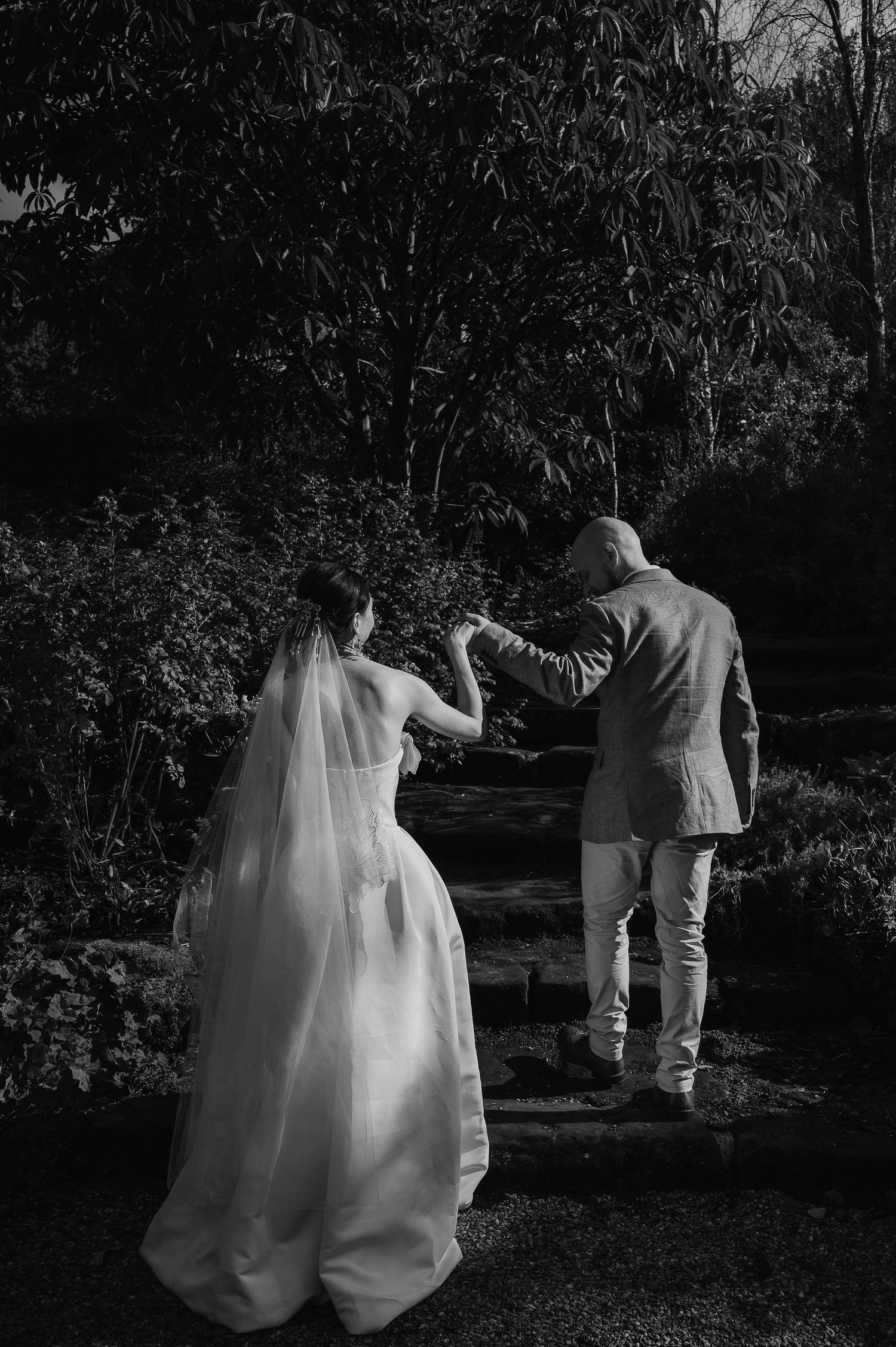 Black and white photo of newlyweds walking up garden steps holding hands.
