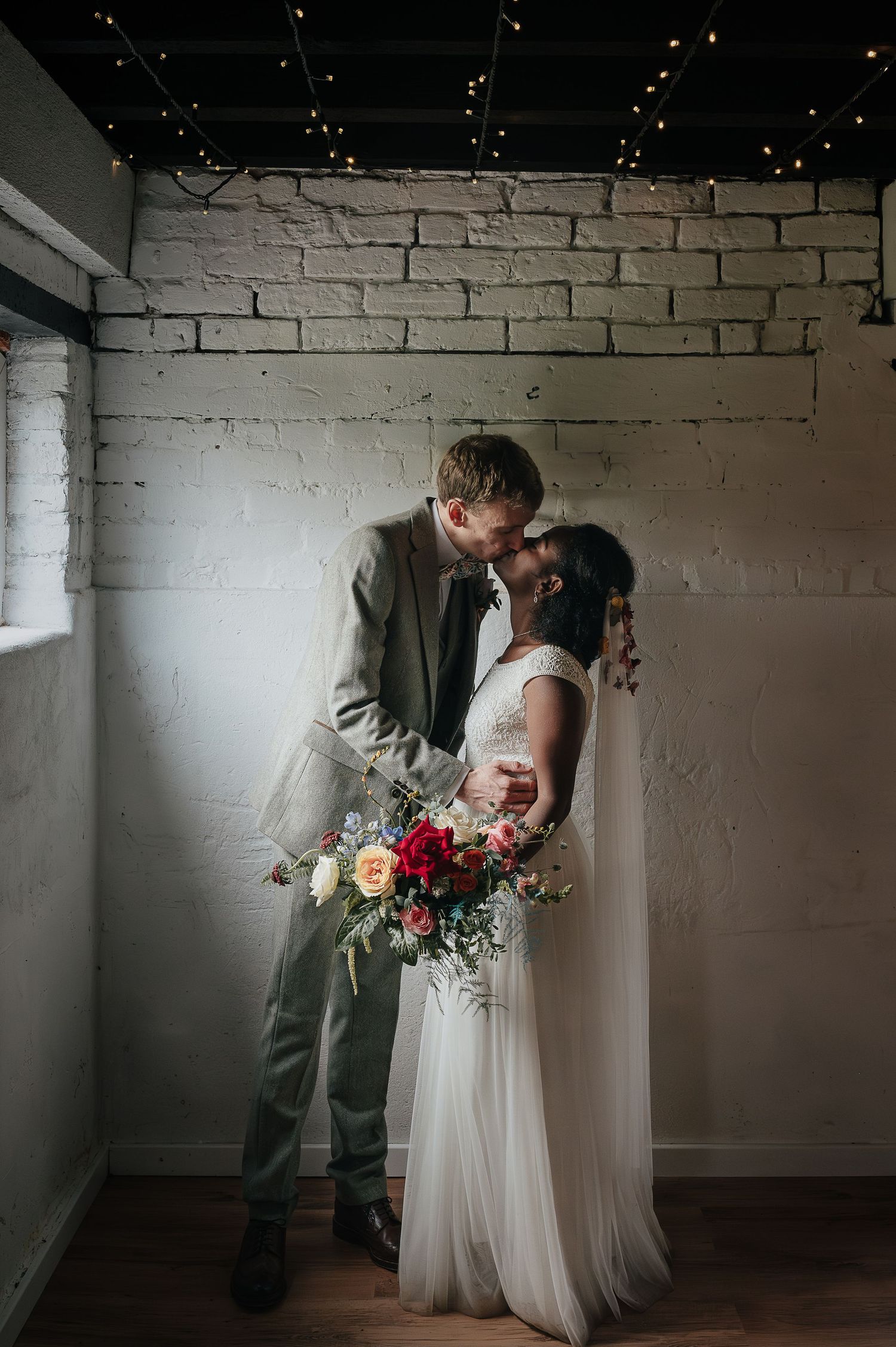 A romantic wedding moment captured in an industrial space with white brick walls and twinkling lights.