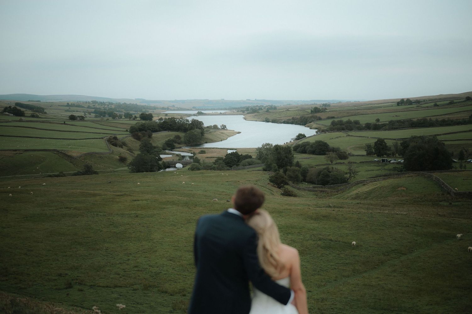 Wedding couple poses against scenic countryside landscape with rolling hills and water views.