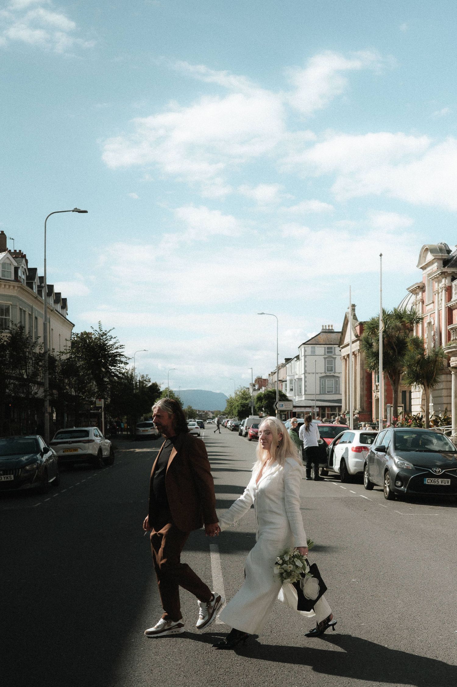 Couple walking down a sunny street lined with parked cars and historic buildings.