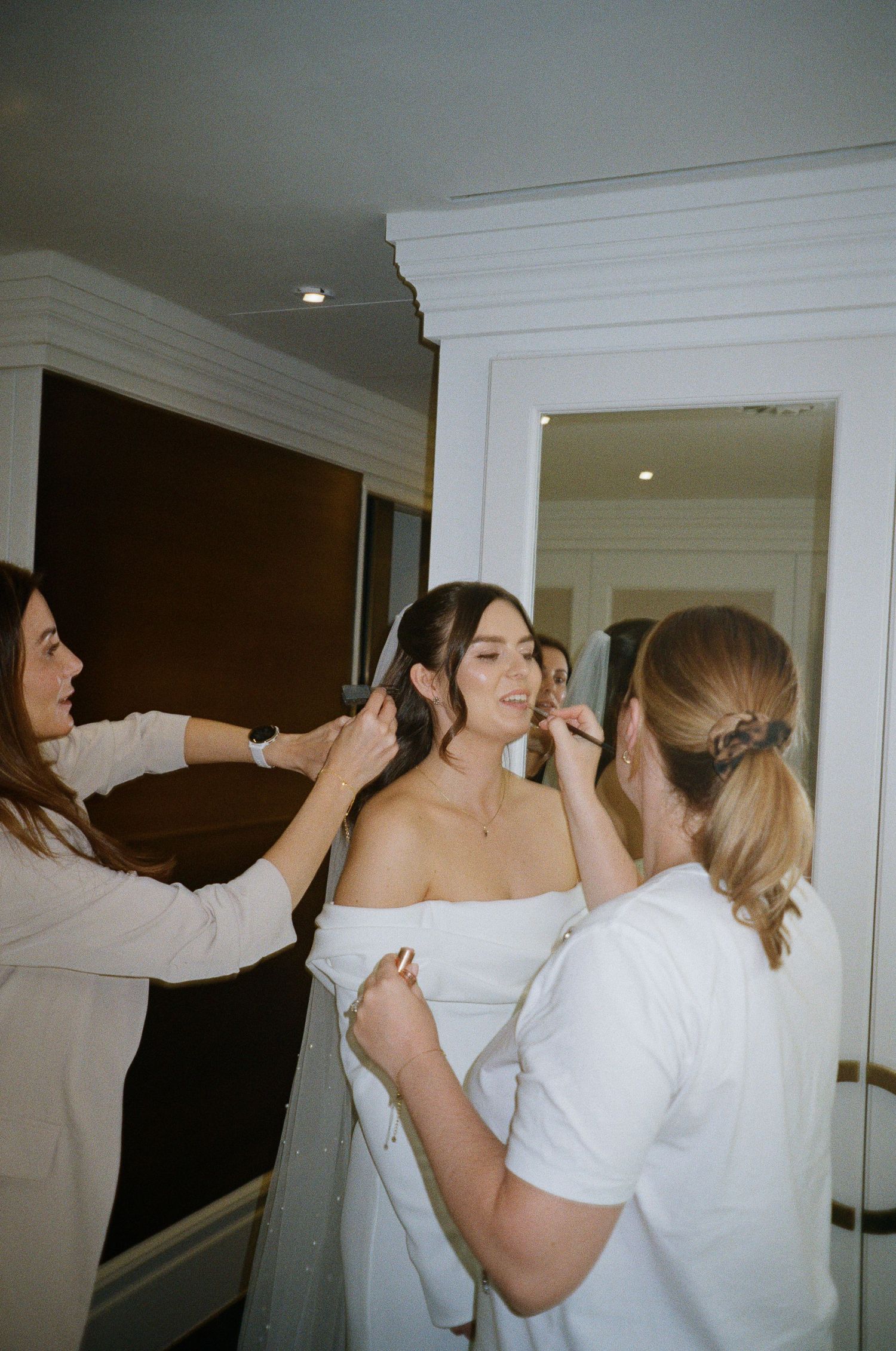Bride getting hair and makeup done by two stylists in a white room before her wedding ceremony.