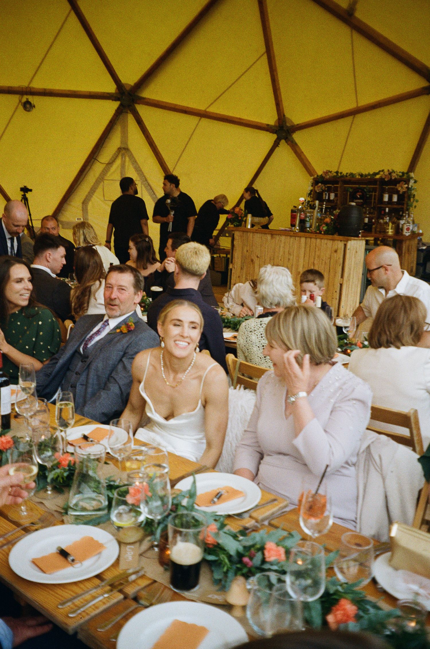 Bride smiling joyfully at wedding reception dinner inside a yellow geodesic dome tent with guests.