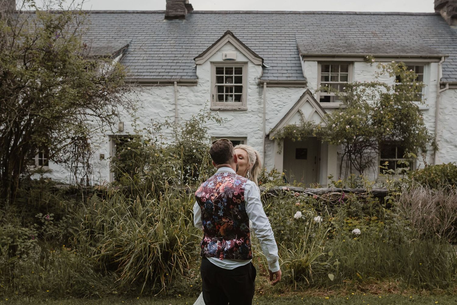 A couple embraces in front of a charming white country house with overgrown garden.