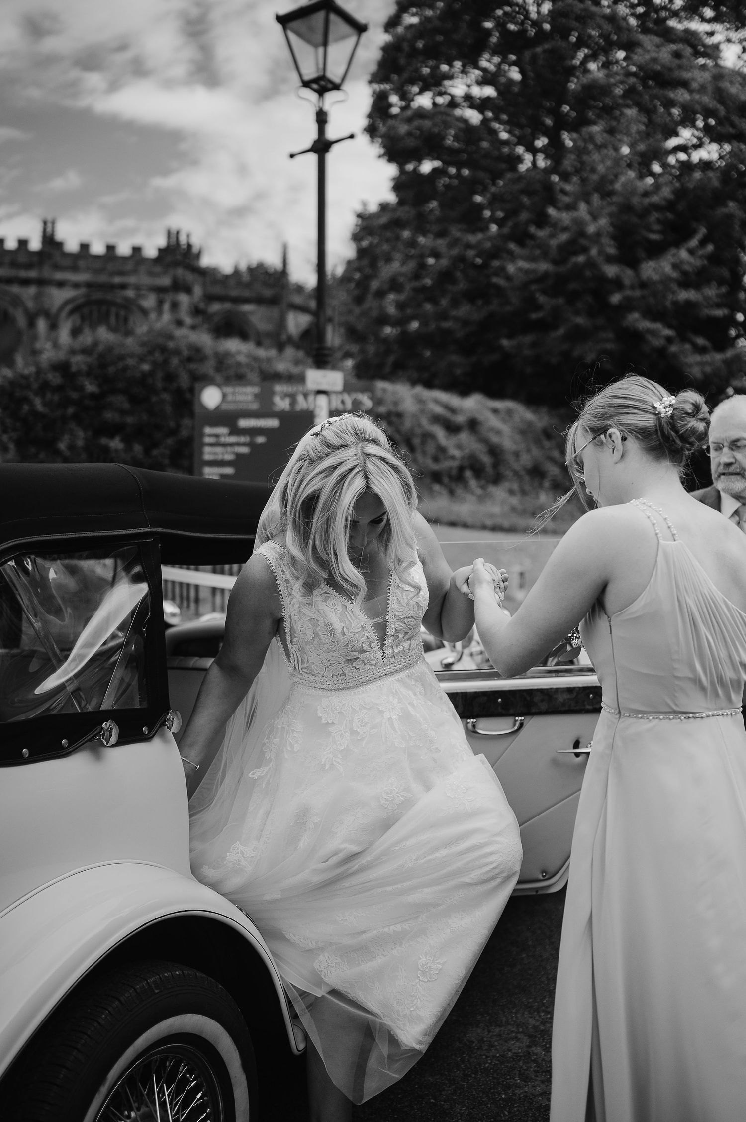 Bride in a lace gown getting assistance entering a vintage car on her wedding day.