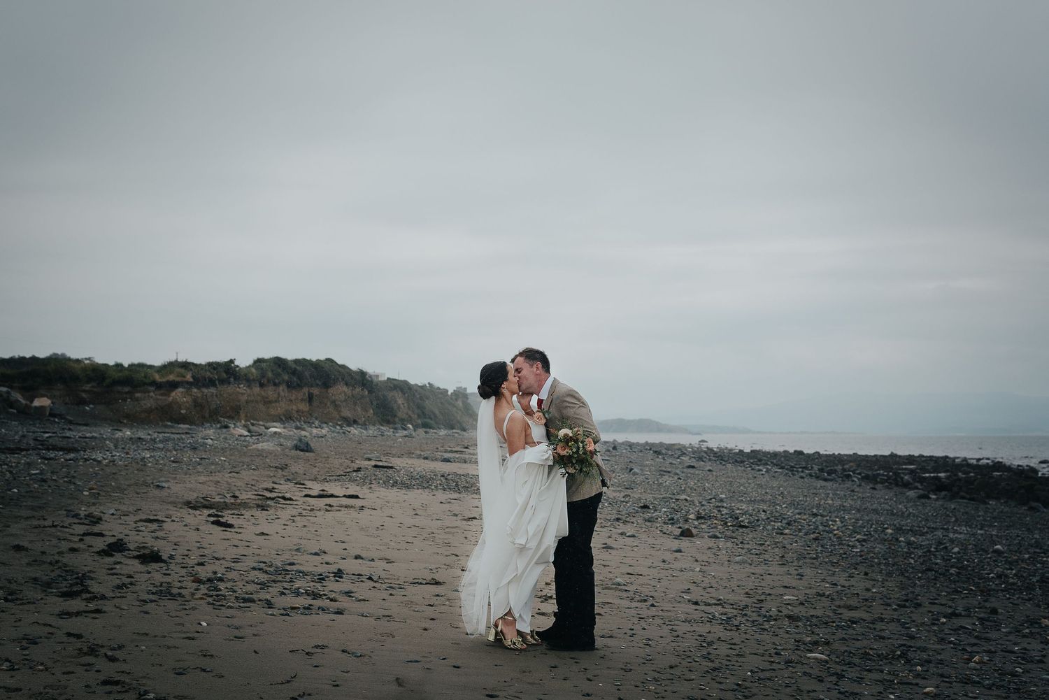 A couple shares an intimate moment on a rocky beach under an overcast sky.