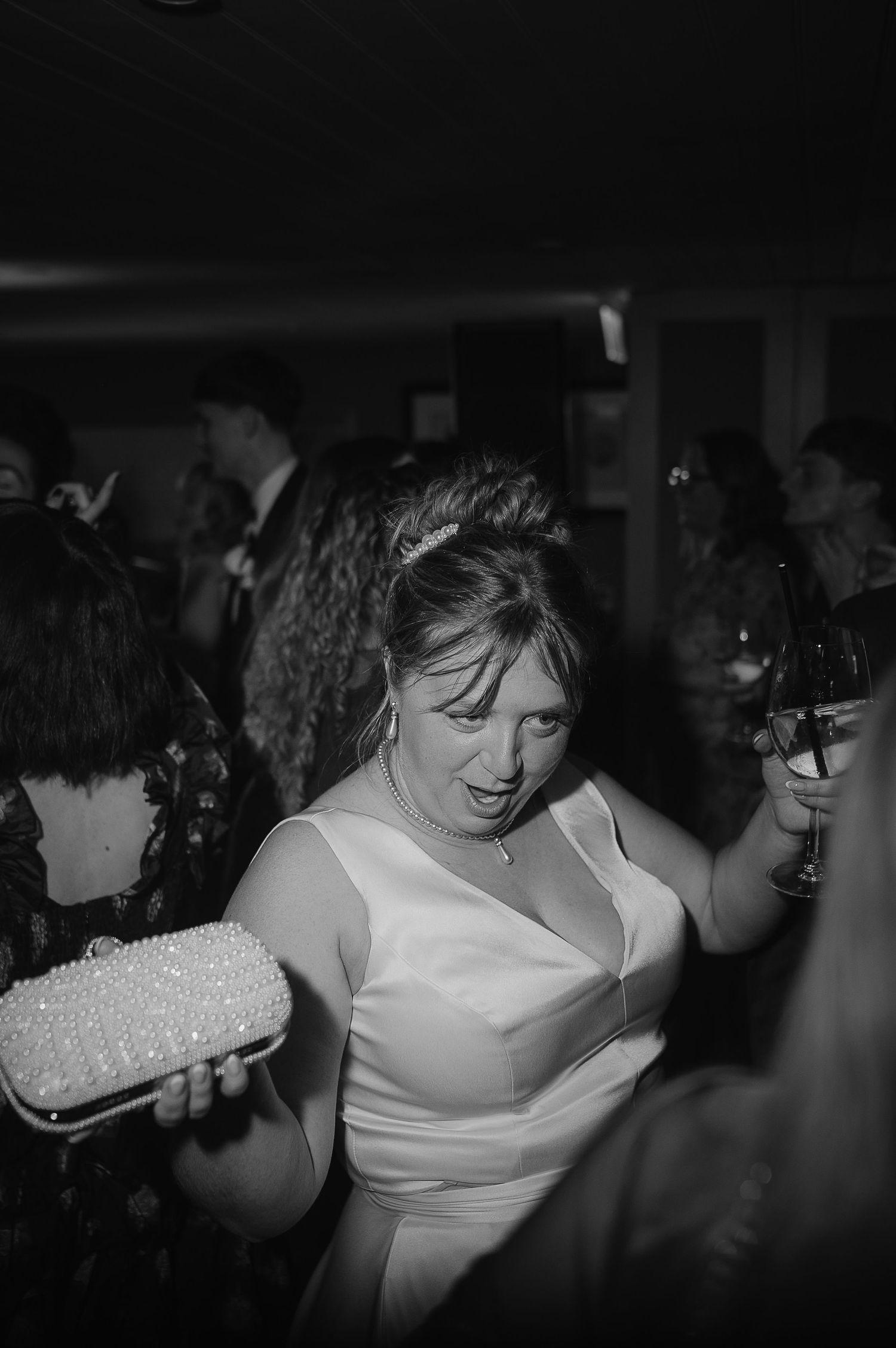 A person wearing a white sleeveless dress holds a beaded purse while dancing at a party in black and white photography.