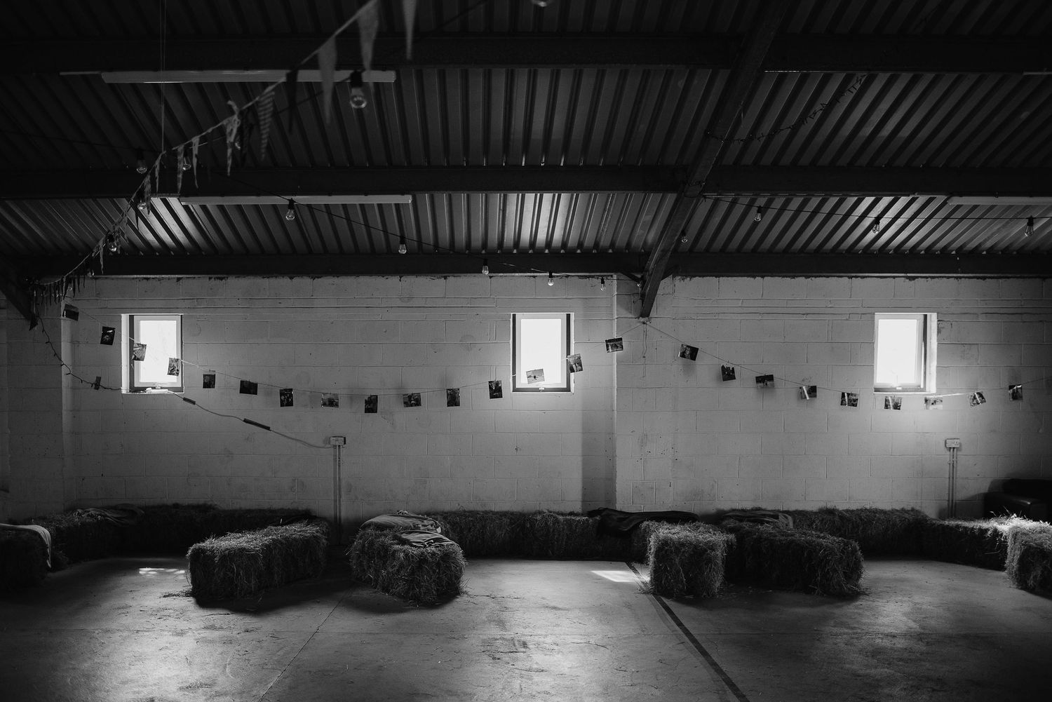 Black and white photo of rustic barn interior with string lights and hay bales.