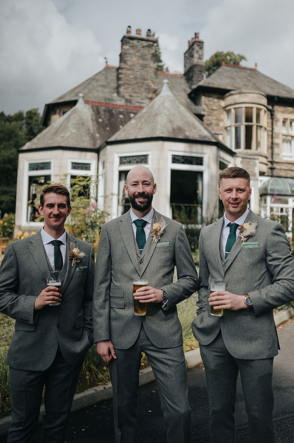 Three groomsmen in matching gray suits holding drinks outside a stone estate.