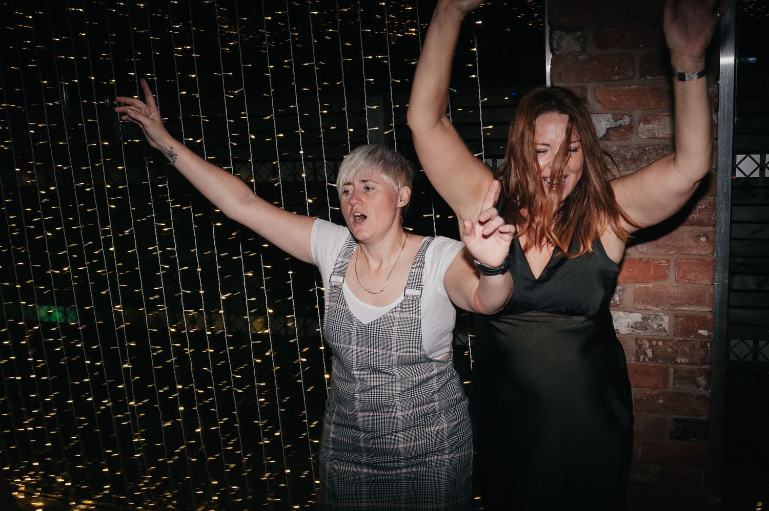 Two wedding guests dancing enthusiastically under twinkling fairy lights against a brick wall.