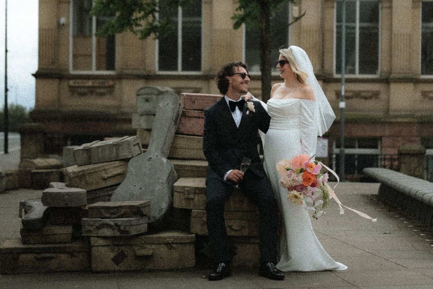 Bride in white gown and groom in black tuxedo pose on stone steps outside a historic building on their wedding day.