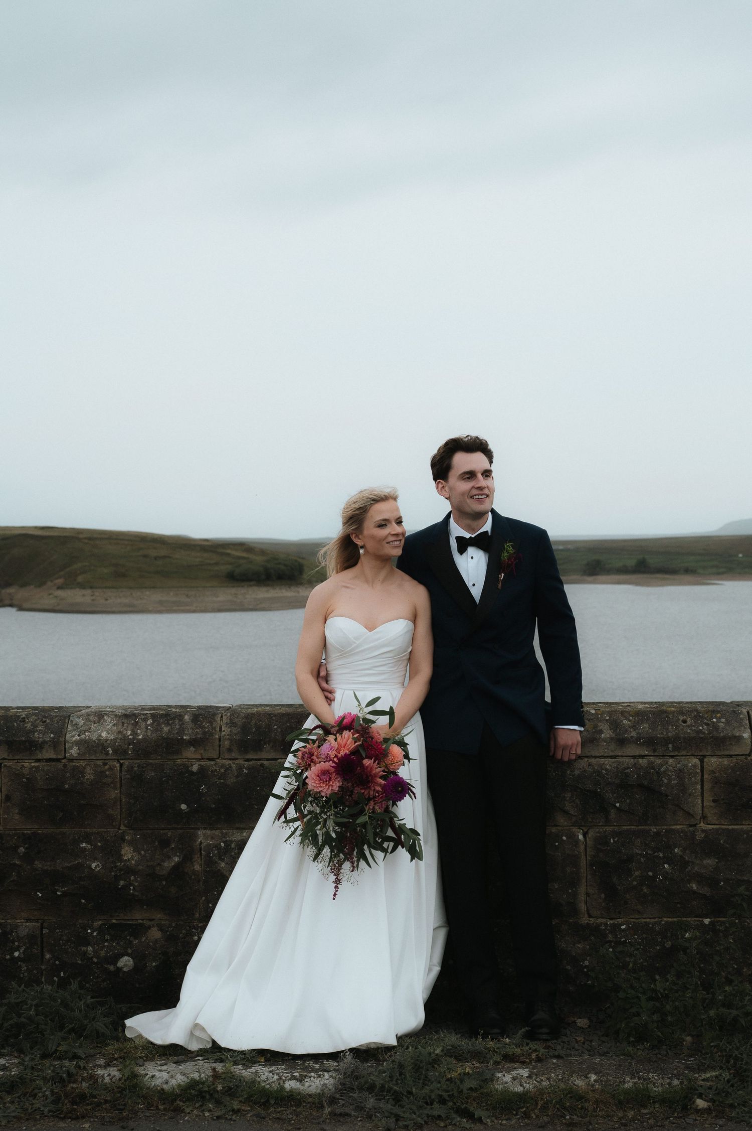 A newlywed couple poses along a stone wall overlooking a coastal waterway with moody skies. Luke Hassall Photography.