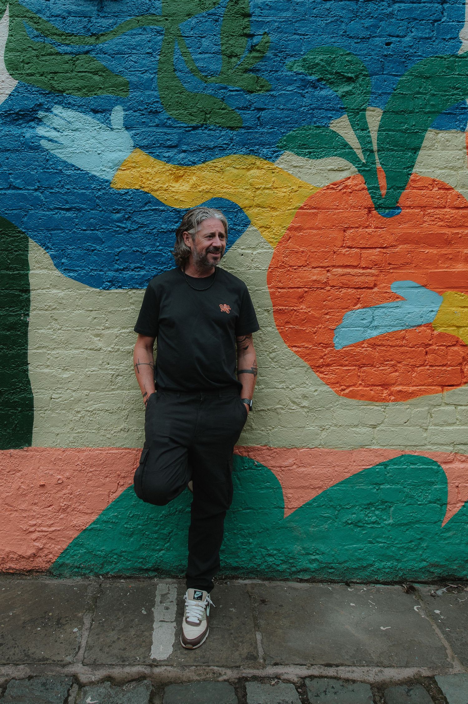 A person in dark clothing poses against a colorful outdoor mural with abstract shapes in blue, orange and green tones.