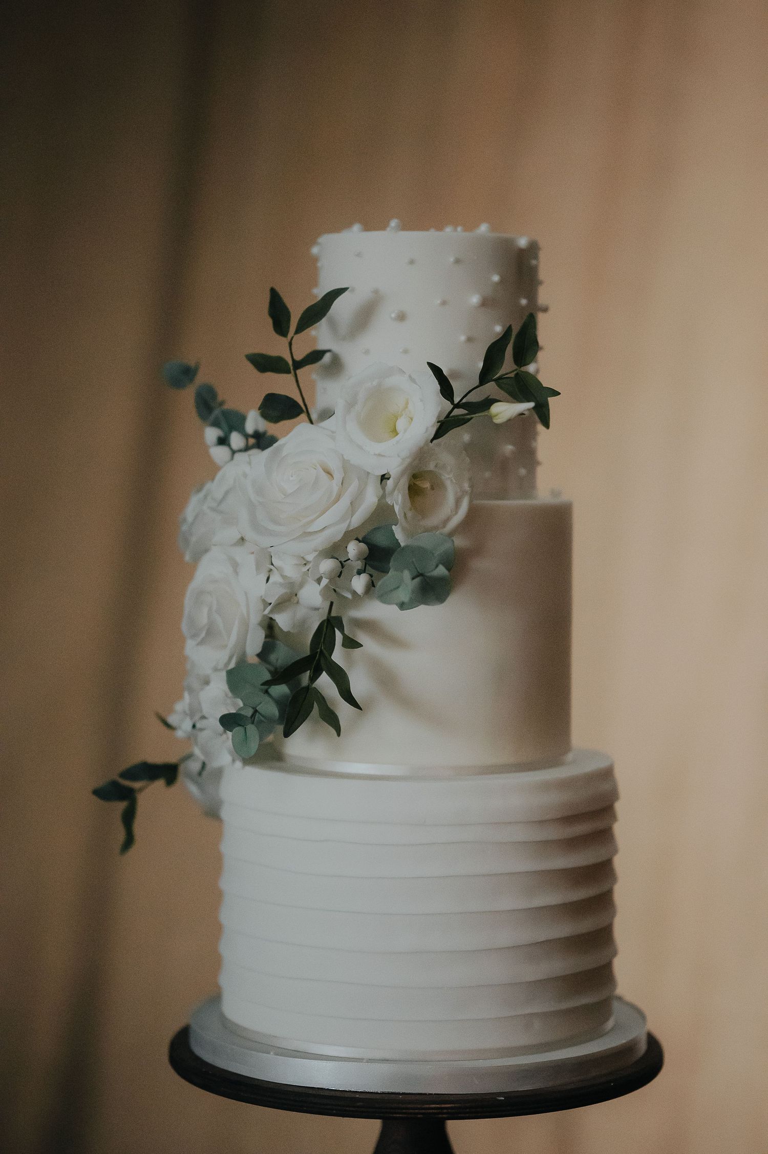 Elegant three-tiered white wedding cake decorated with roses and greenery against a beige background.