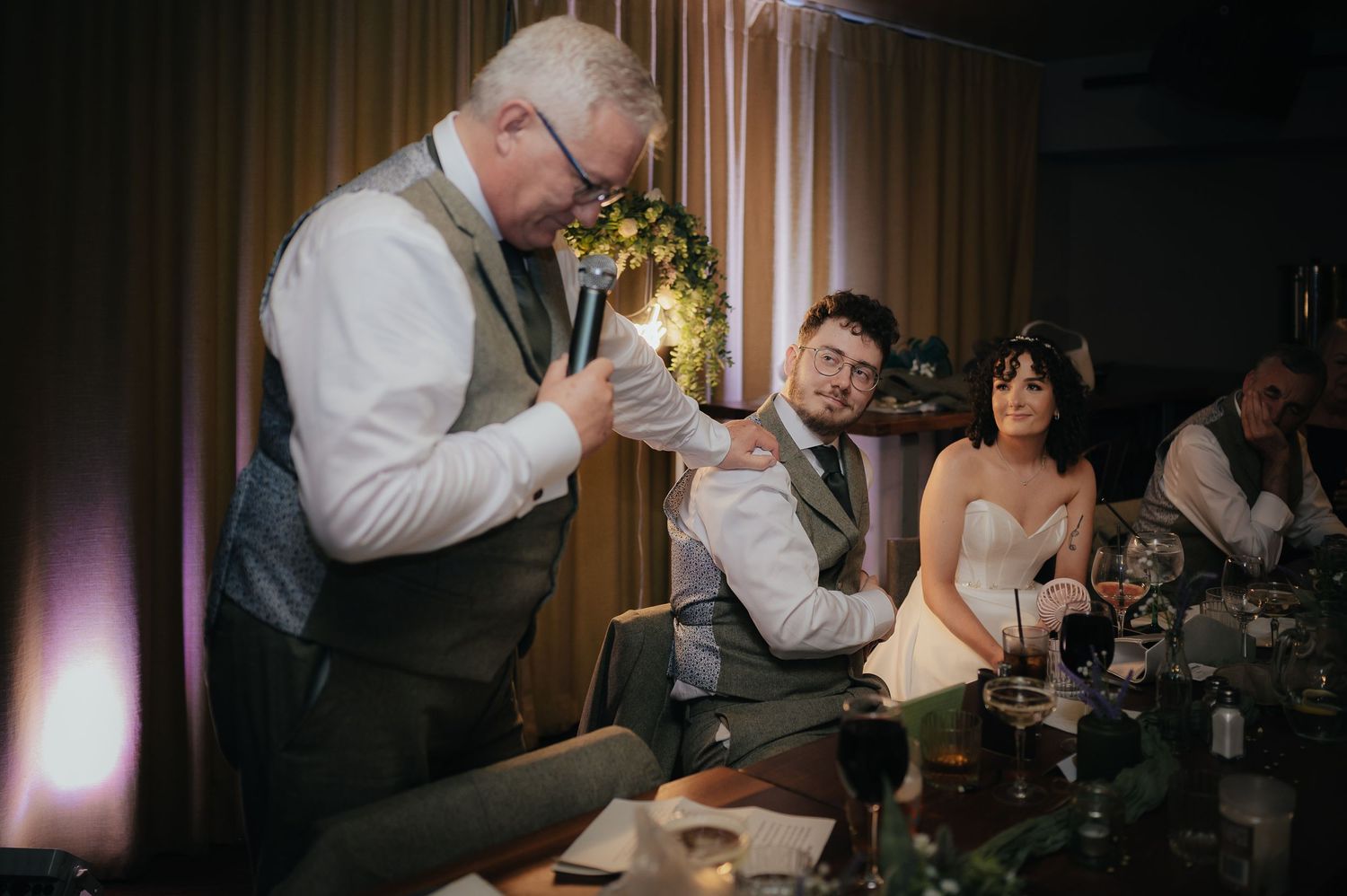 Wedding party shares drinks and laughter at reception table with decorative lighting.