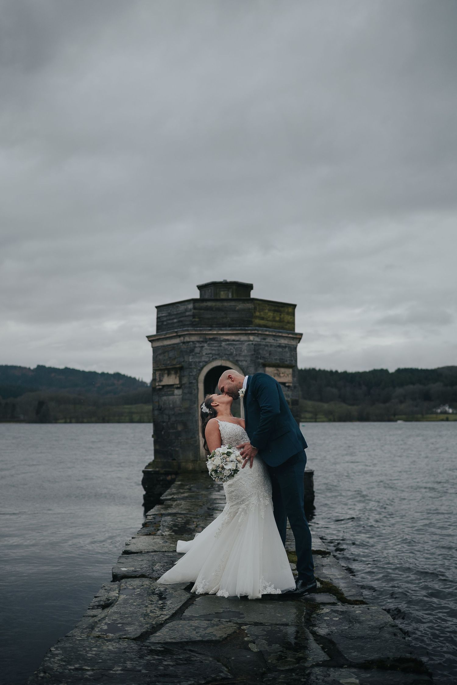 A couple shares an intimate moment on a stone pier by a moody lake under overcast skies.