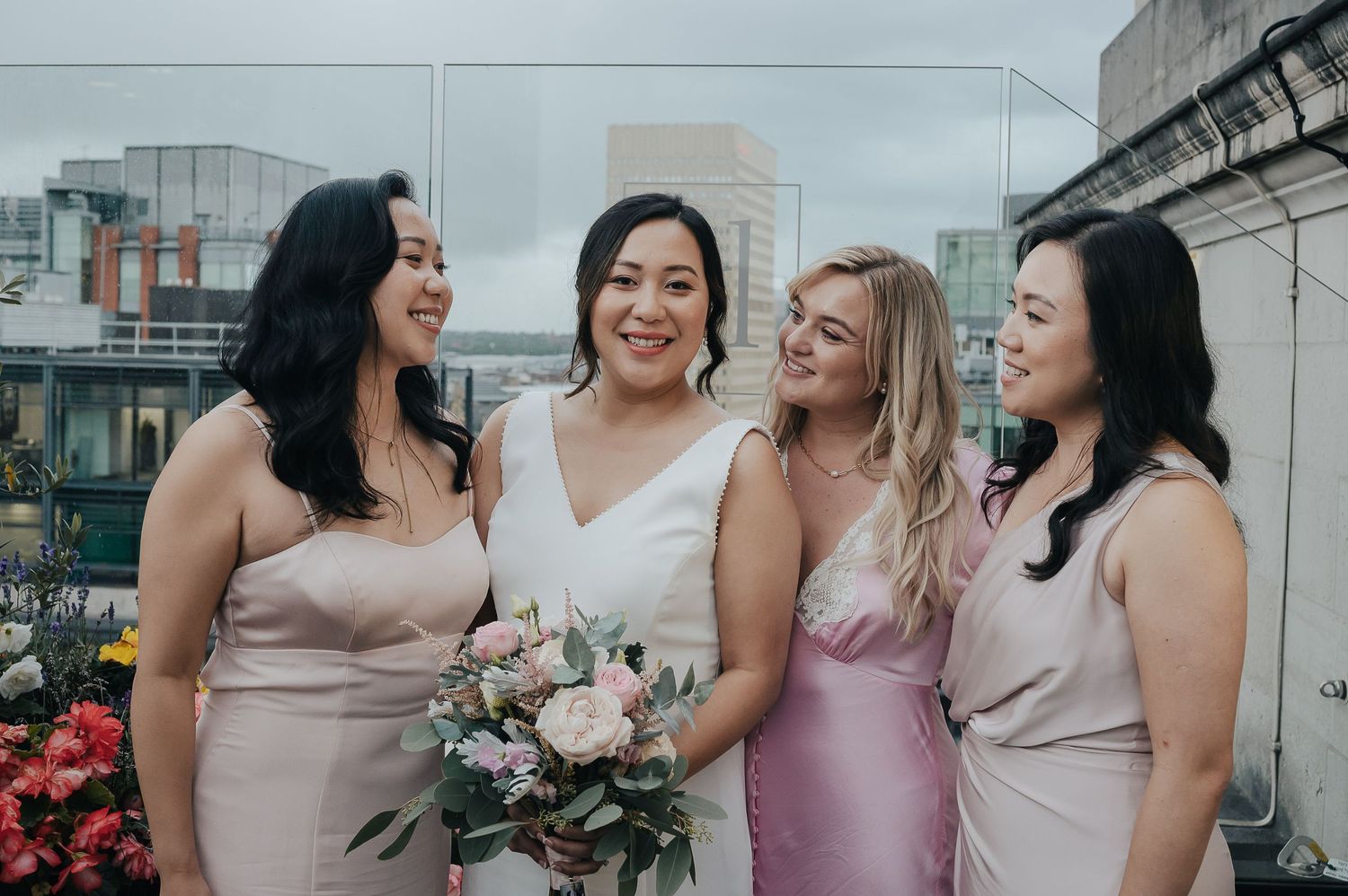 Bridesmaids in pastel dresses pose together on a rooftop with flowers.