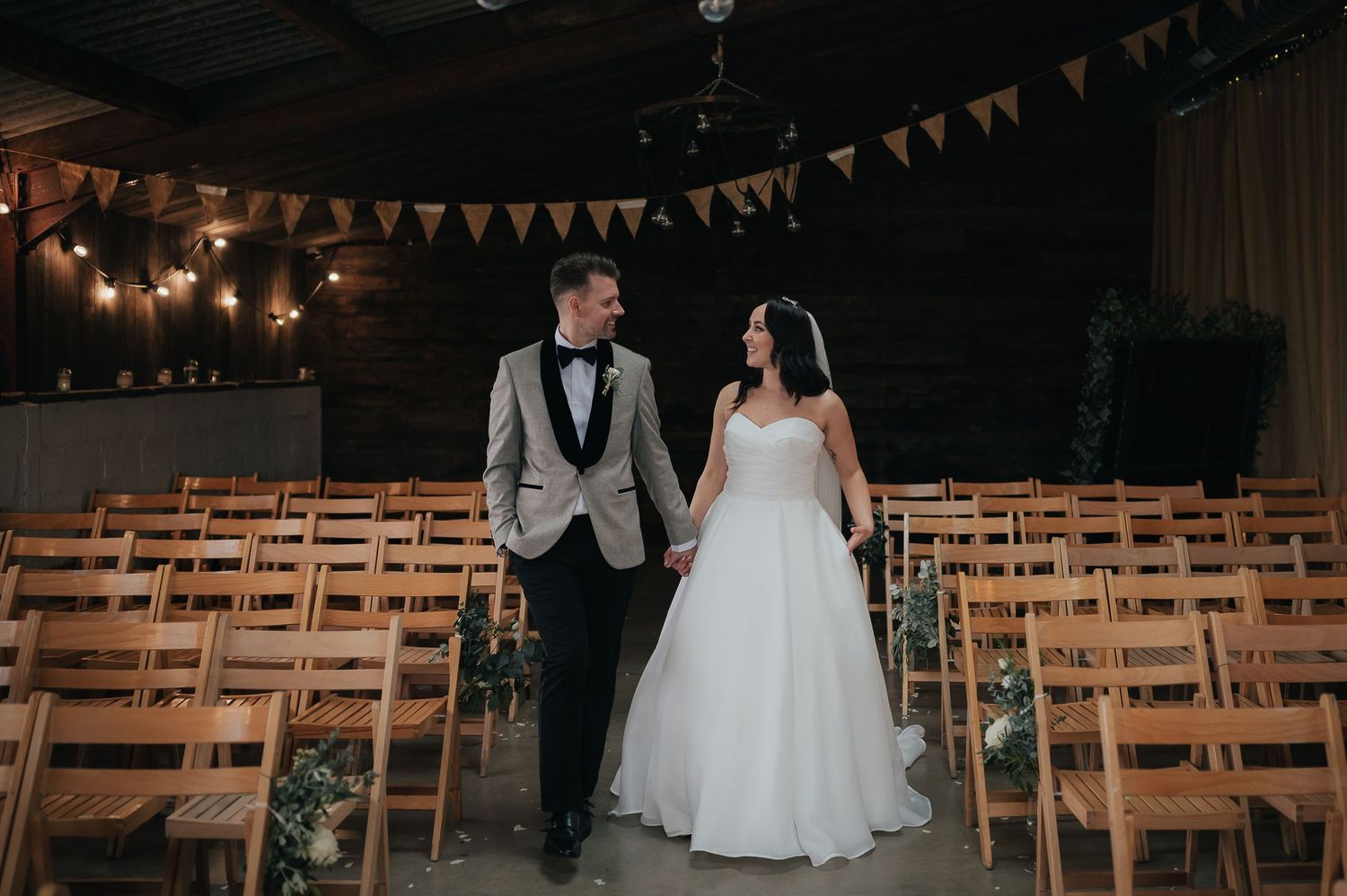 A couple stands in a rustic wedding venue with wooden chairs and string lights hanging overhead.