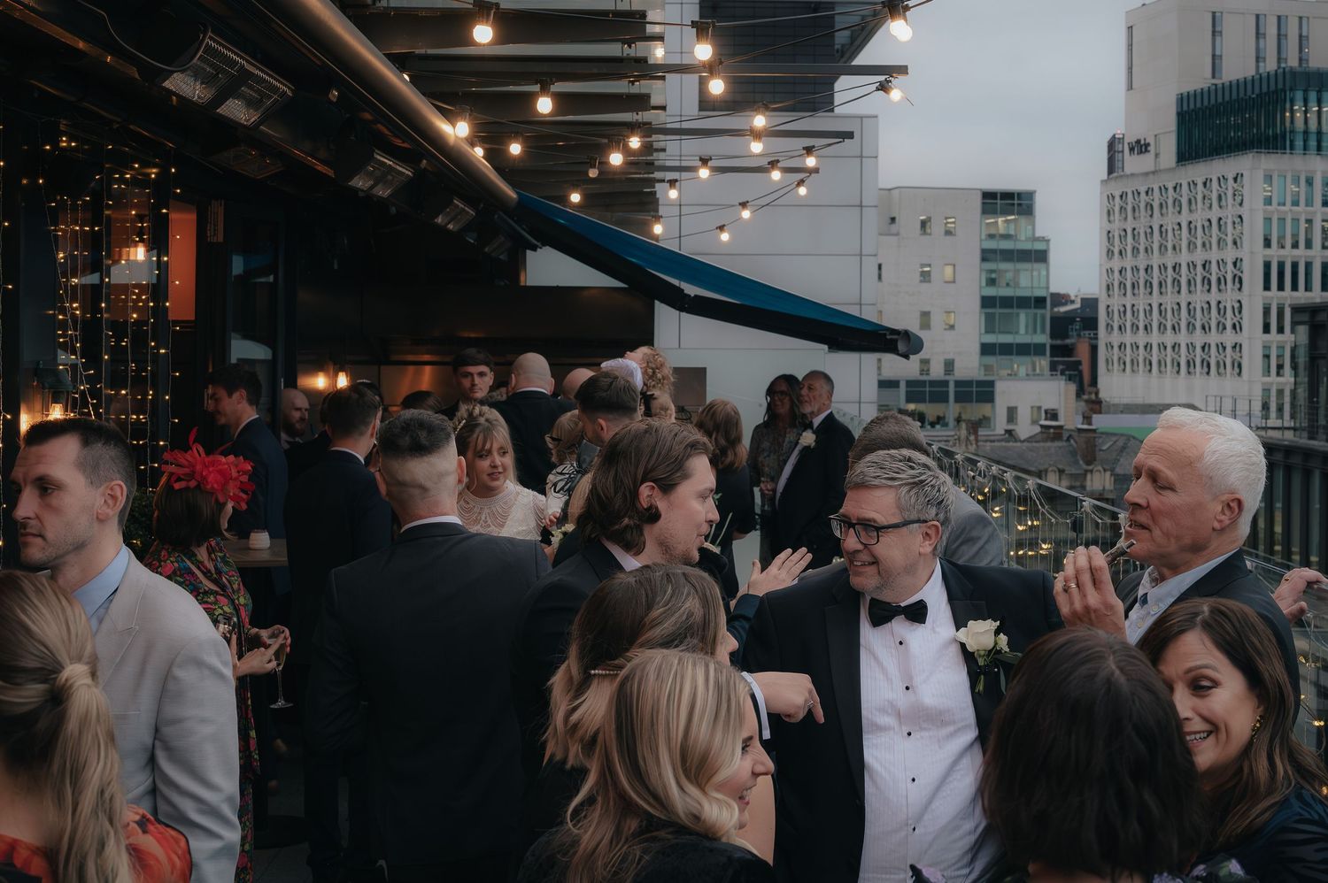 Crowded rooftop party with guests in formal attire mingling under string lights in an urban setting.