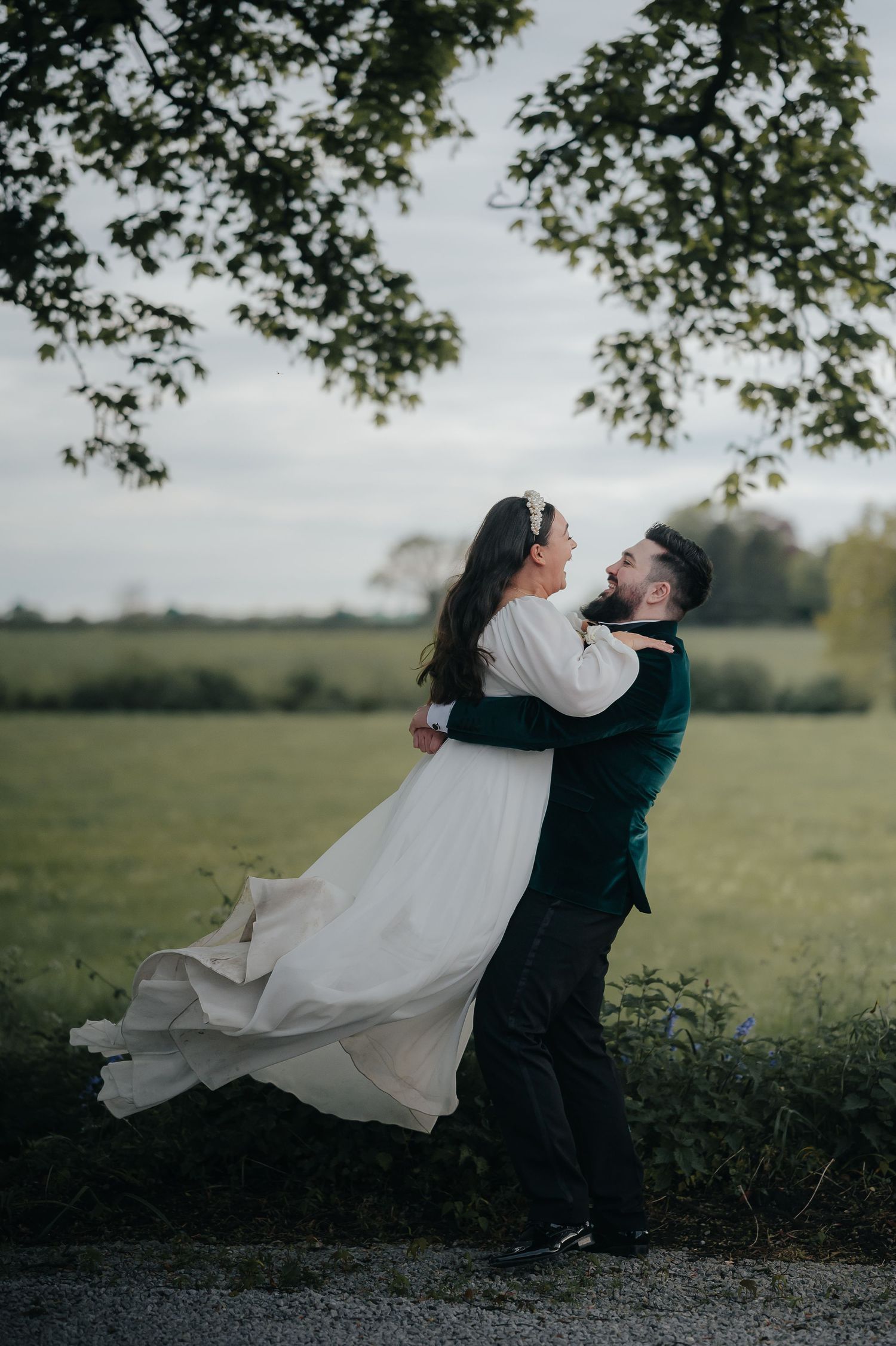 Couple in wedding attire embracing under tree branches in a field.