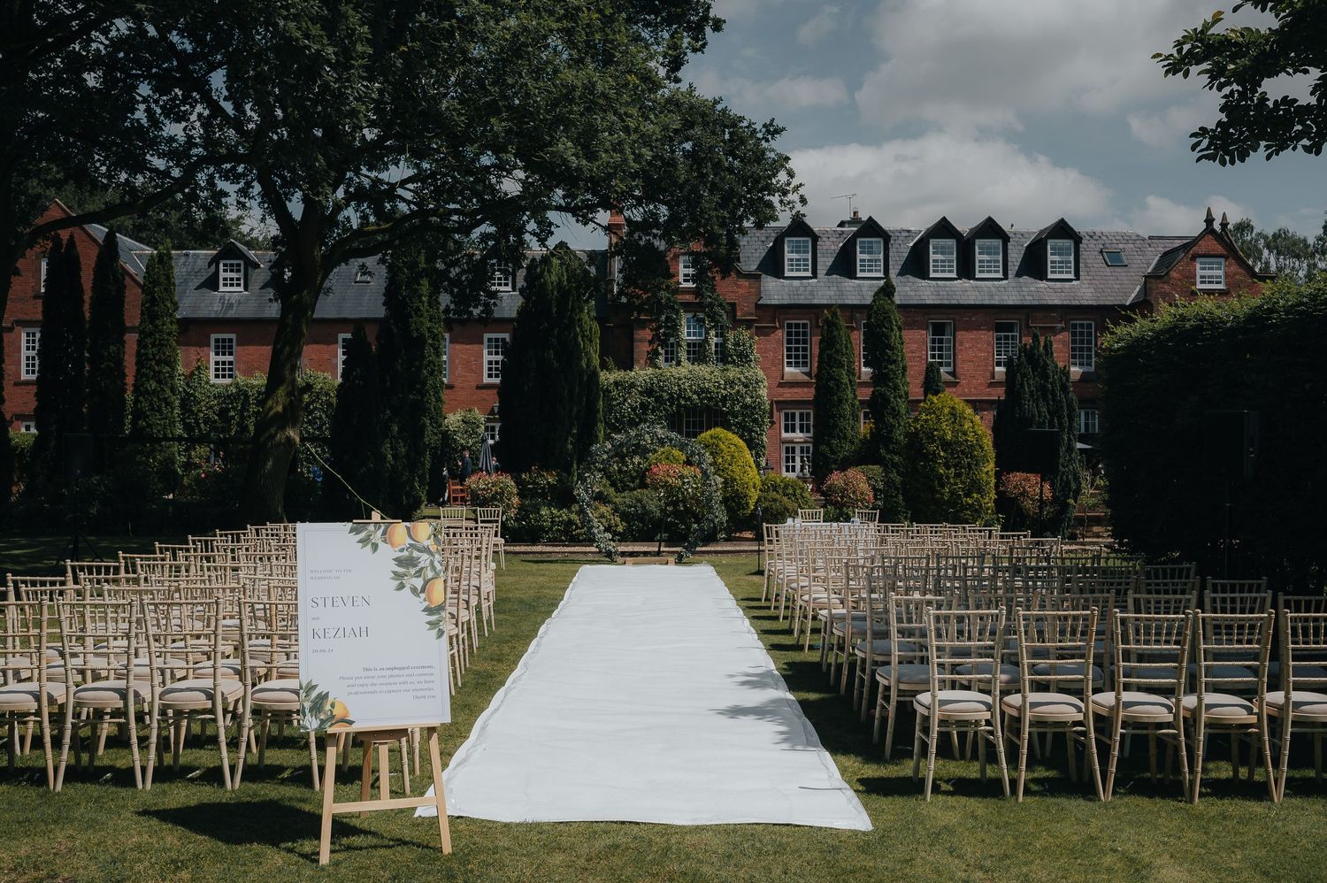 Outdoor wedding ceremony setup with white aisle runner and wooden chairs against brick building backdrop.
