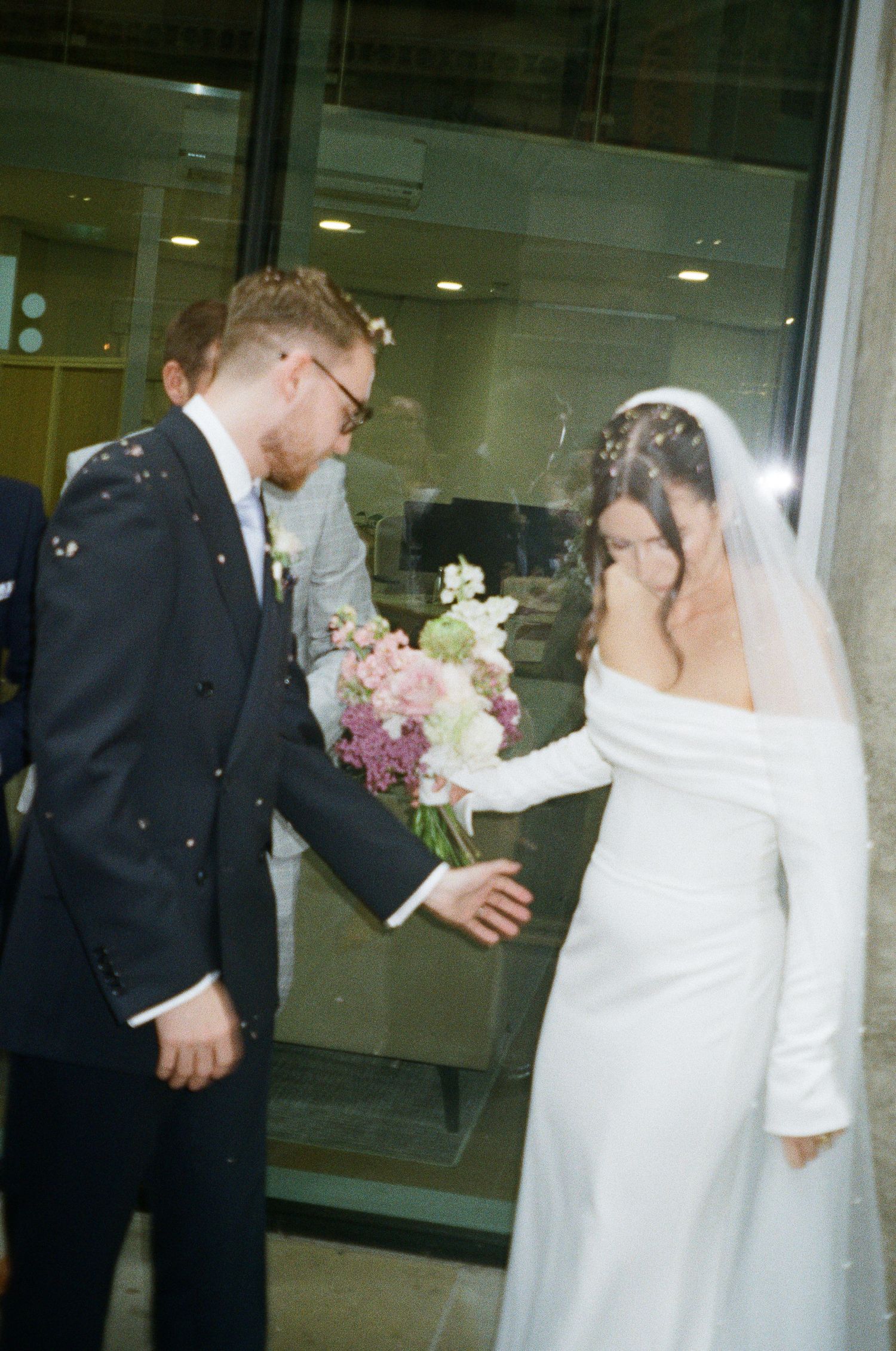 Bride in white off-shoulder gown and veil holds bouquet with groom in dark suit as confetti falls outside.