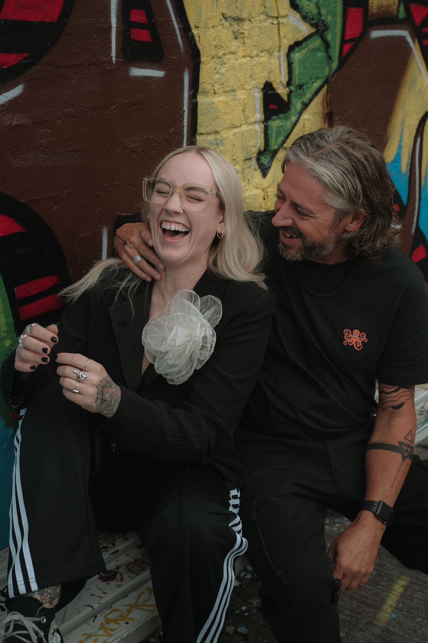Two people in black clothing share a joyful moment of laughter against a brick wall background.
