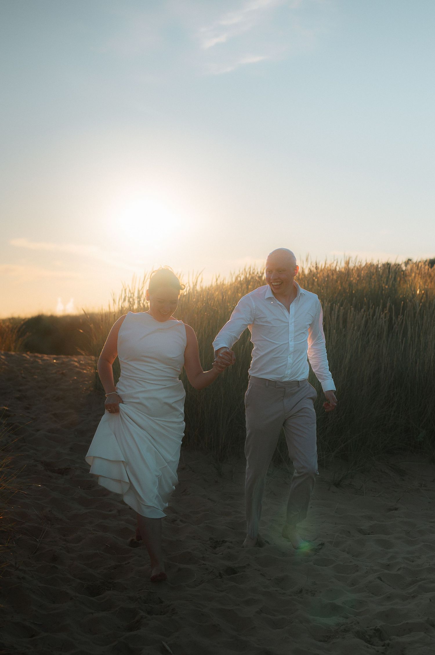 A couple in white attire dances together on a beach at sunset with tall grass in the background.