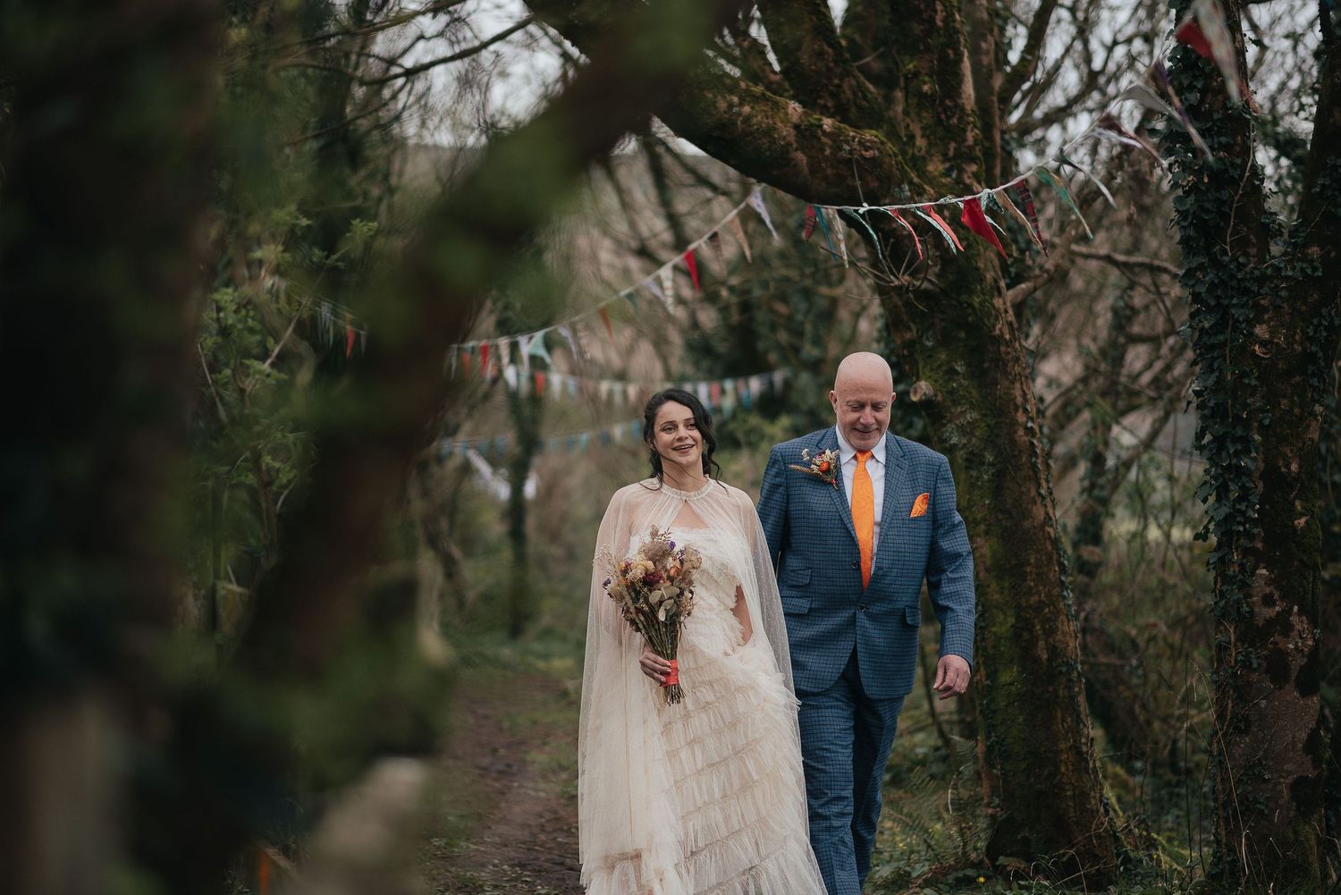 A couple walks down a woodland path in wedding attire with fairy lights strung above.