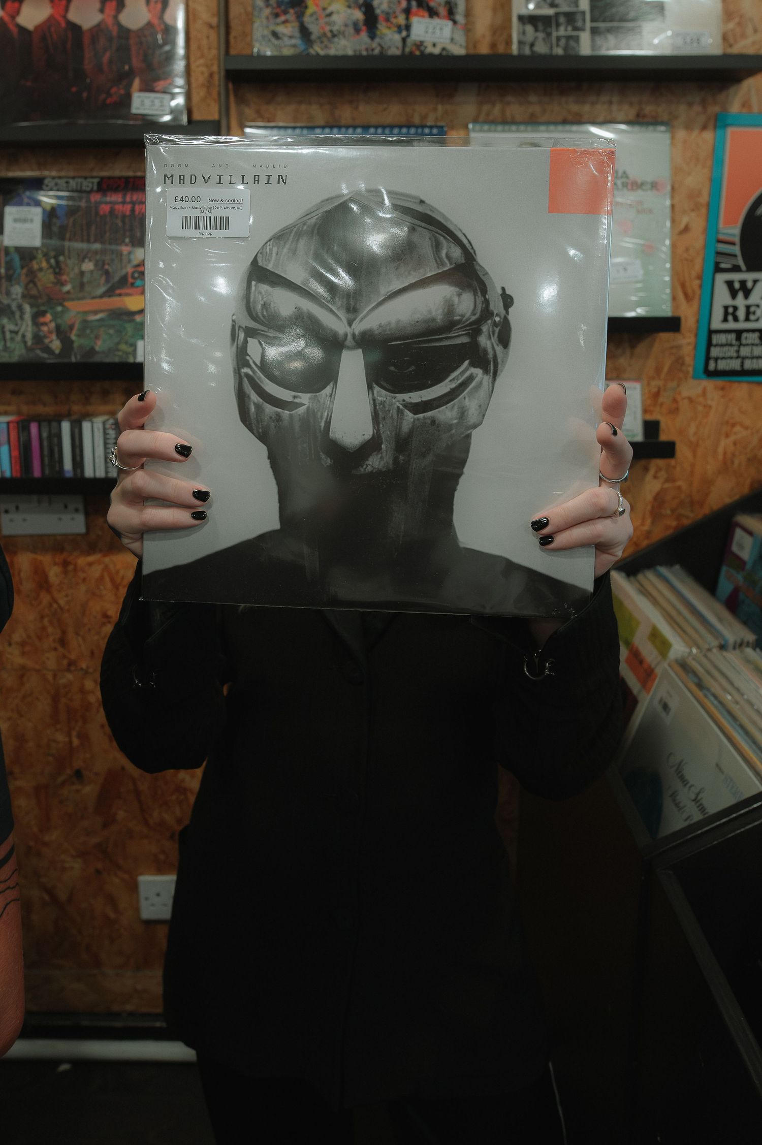Hands hold up a vinyl record sleeve showing a metallic mask design against a wooden wall background.